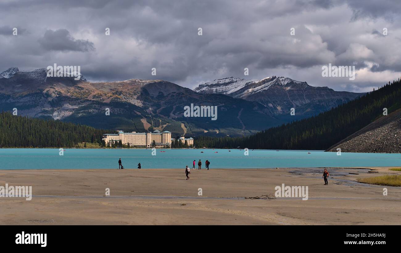 Touristen genießen den Spaziergang am Ufer des Lake Louise mit türkisfarbenem Wasser und Hotelkomplex im Hintergrund im Banff National Park, Kanada. Stockfoto