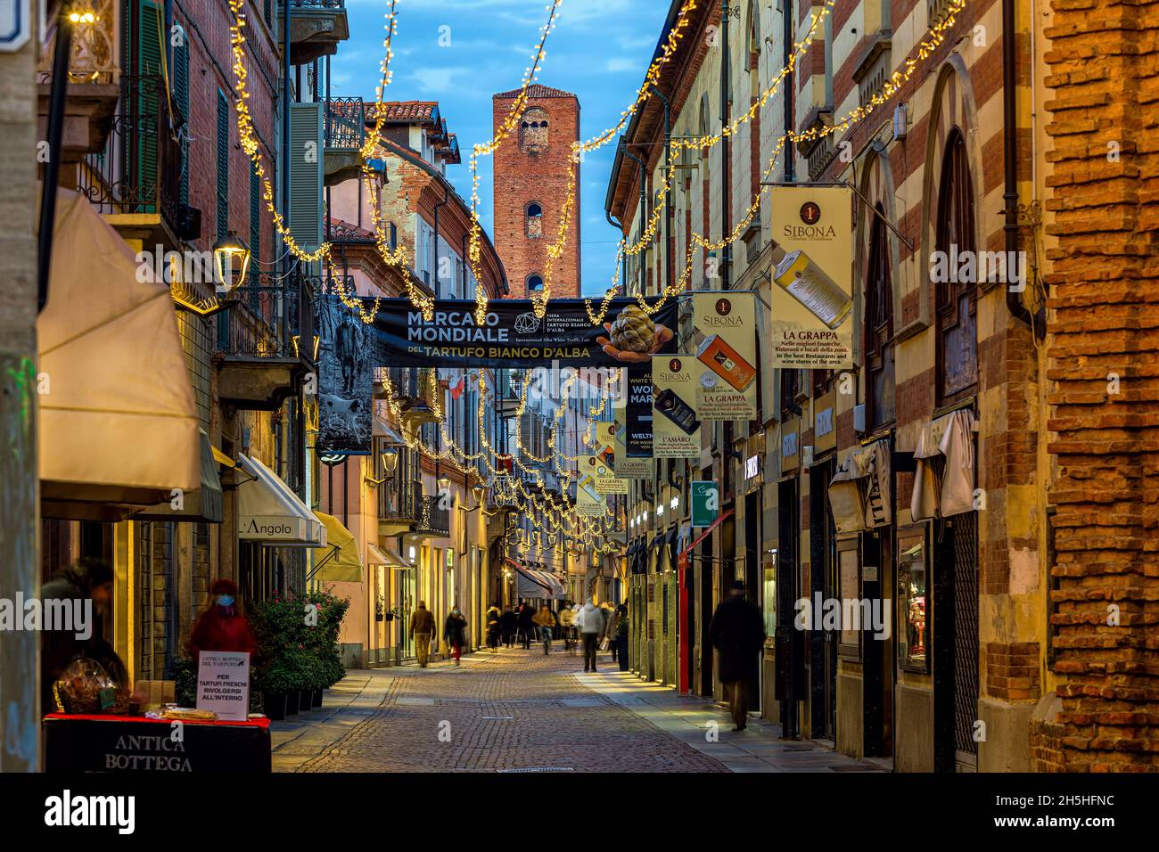 Menschen, die abends in der Altstadt von Alba, Piemont, Norditalien, auf der von Weihnachtslichtern beleuchteten zentralen Straße spazieren gehen. Stockfoto