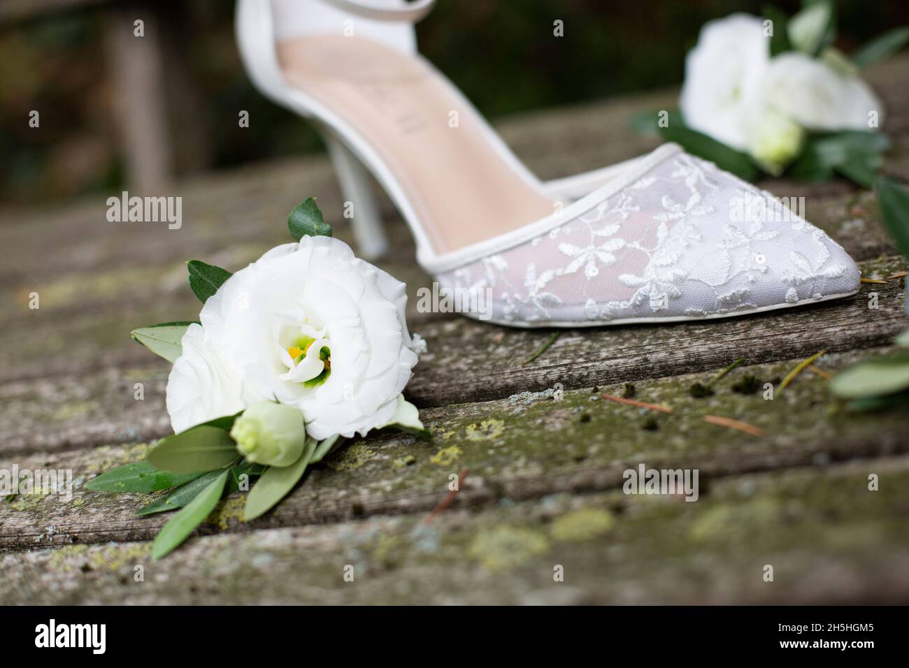 Detail eines einzigen weißen Hochzeitsschuhs mit Stickerei und Absatz, der auf alten ruinierten Holzplanken platziert und von weißen Blumen mit ol umgeben ist Stockfoto