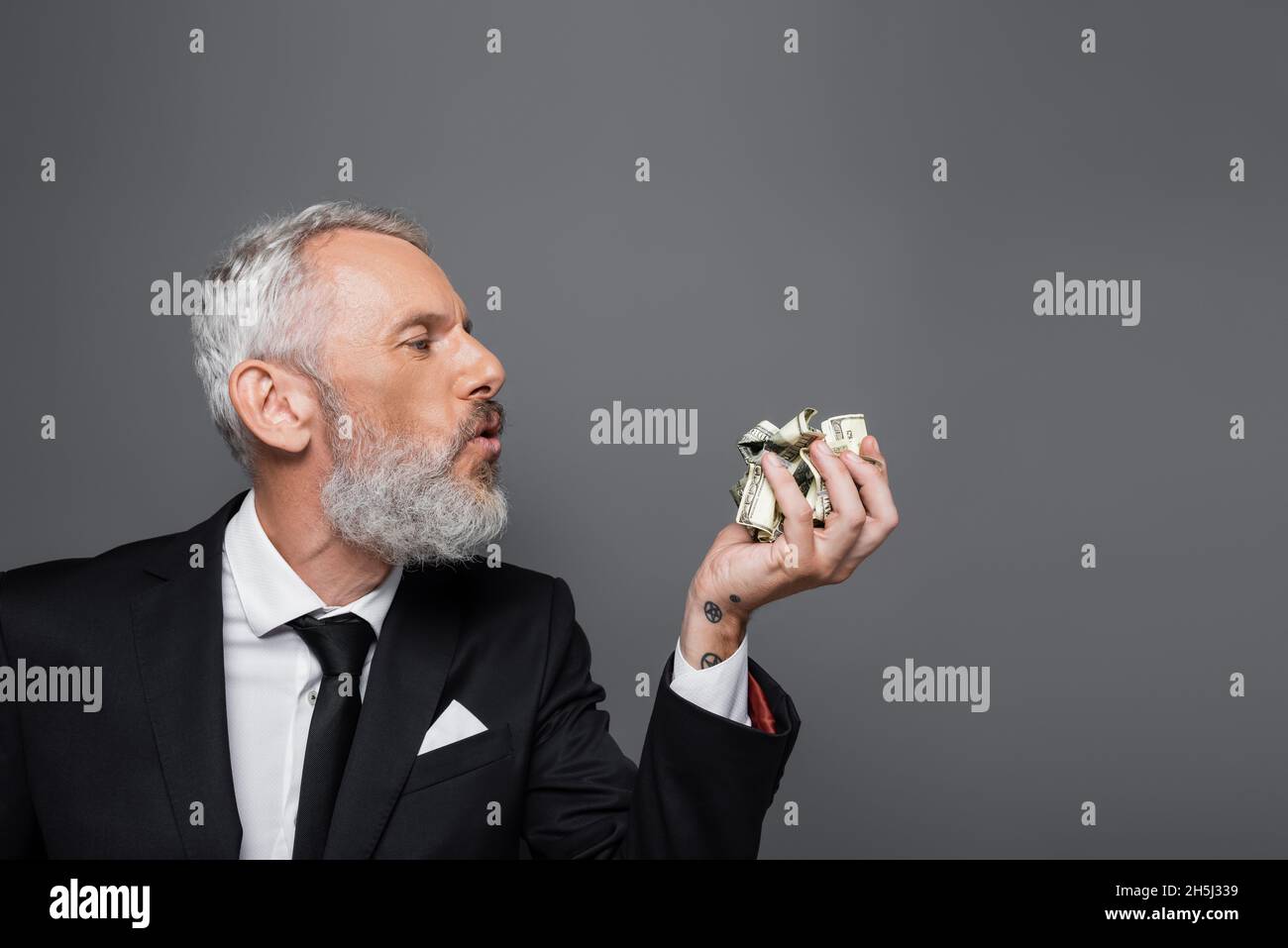 Seitenansicht eines bärtigen Geschäftsmannes mittleren Alters, der auf zerknitterte Dollar-Banknoten bläst, die isoliert auf Grau liegen Stockfoto