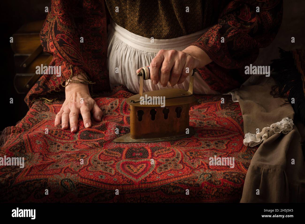 Alte Frau im viktorianischen Kleid, die mit einem antiken Holzkohleisen auf einem Brett arbeitet Stockfoto