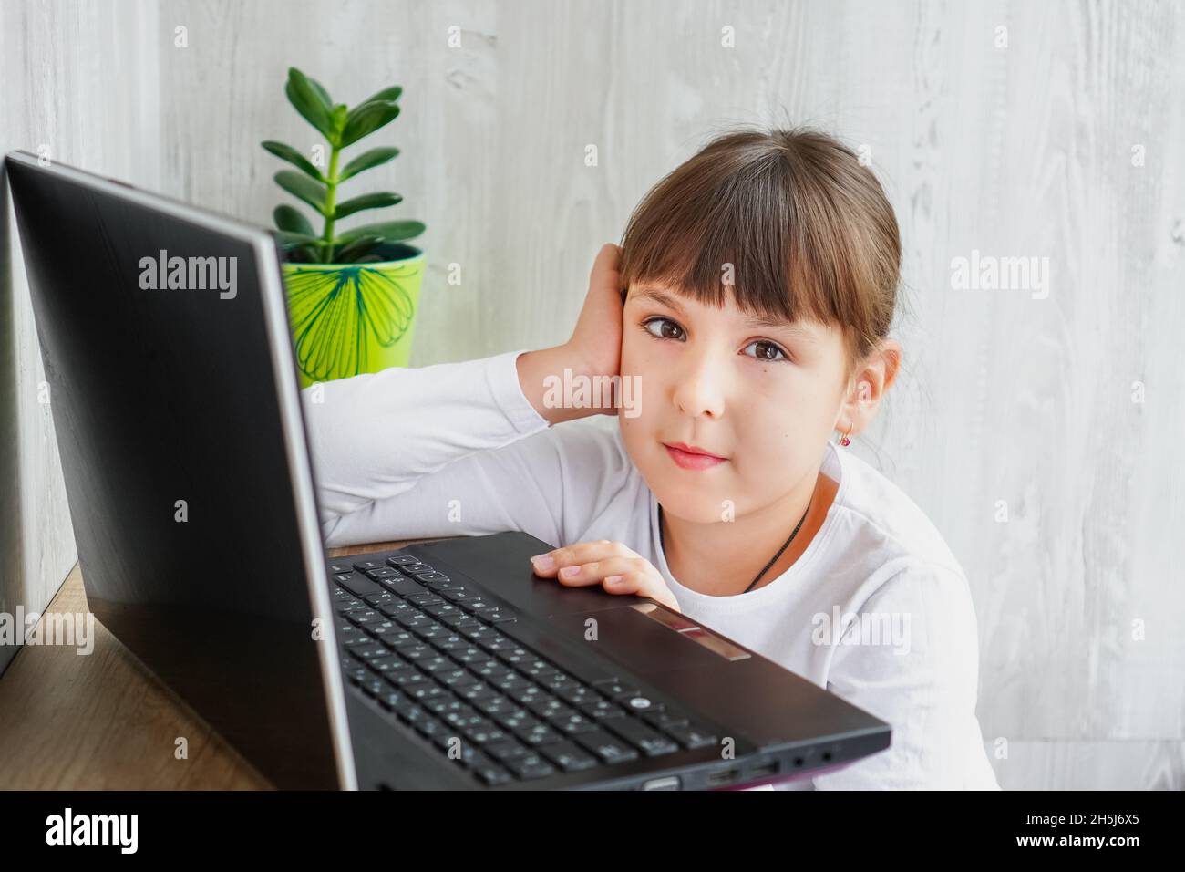 Innenaufnahme eines niedlichen dunkelhaarigen weiblichen Kindes, das am Tisch neben dem Notebook sitzt und die Kamera mit einer Hand im Gesicht betrachtet Stockfoto