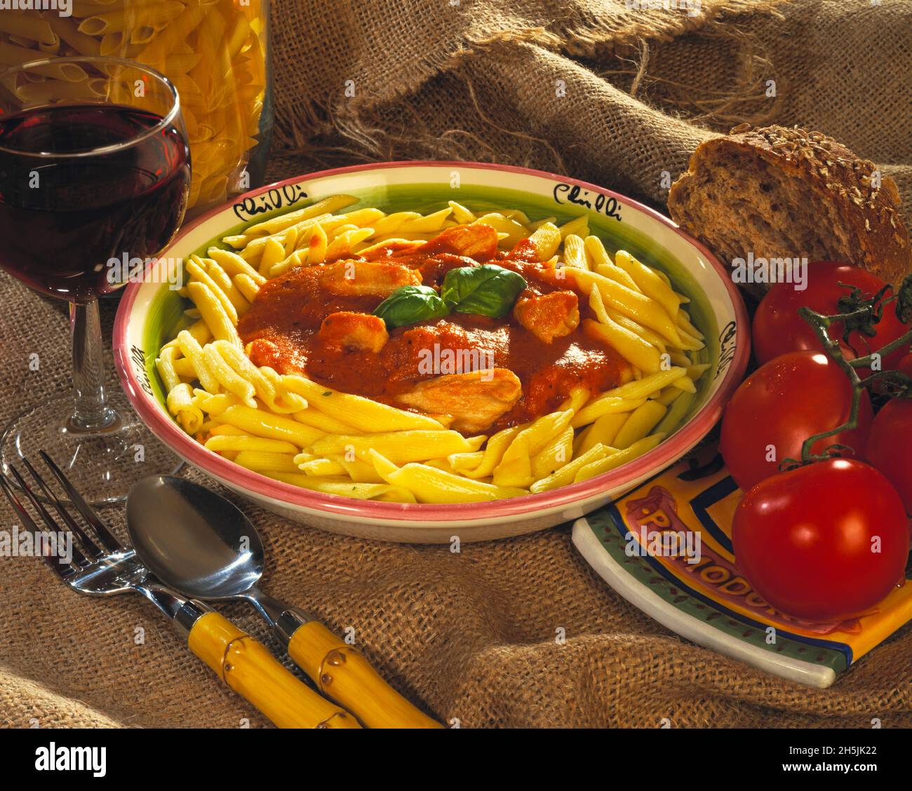 Huhn in einer Tomatensauce italienischen Stil mit gekochter Pasta Bambusgriff Gabel & Löffel Rotwein in Glas ganze Tomaten Getreidebrot hessischen Hintergrund Stockfoto
