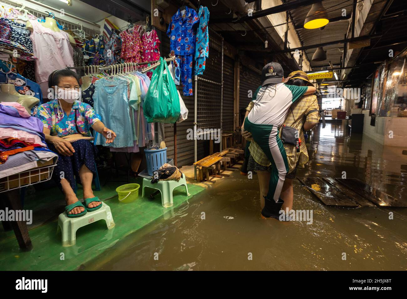 Eine Frau trägt ihren Sohn, während sie durch die Flut geht, Auf der Seite sitzt ein Ladenbesitzer neben ihrem Laden und hebt ihre Füße über das überflutete Wasser im Wang lang Markt.der Chao Phraya Fluss ist überflutet, beeinflusst durch starke Regenfälle, die Überschwemmungen im Wang lang Markt und in vielen niedrig gelegenen Gebieten entlang des Chao Phraya Flusses verursachen. (Foto von Phobthum Yingpaiboonsuk / SOPA Images/Sipa USA) Stockfoto