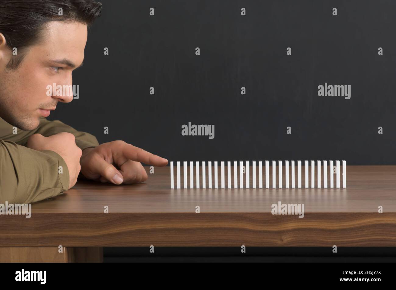 Nahaufnahme eines Mannes, der sich darauf vorbereitet, den ersten Domino in einer Reihe von Dominosteinen auf einen Tisch zu schieben, um das Ergebnis vorwegzunehmen; Deutschland Stockfoto