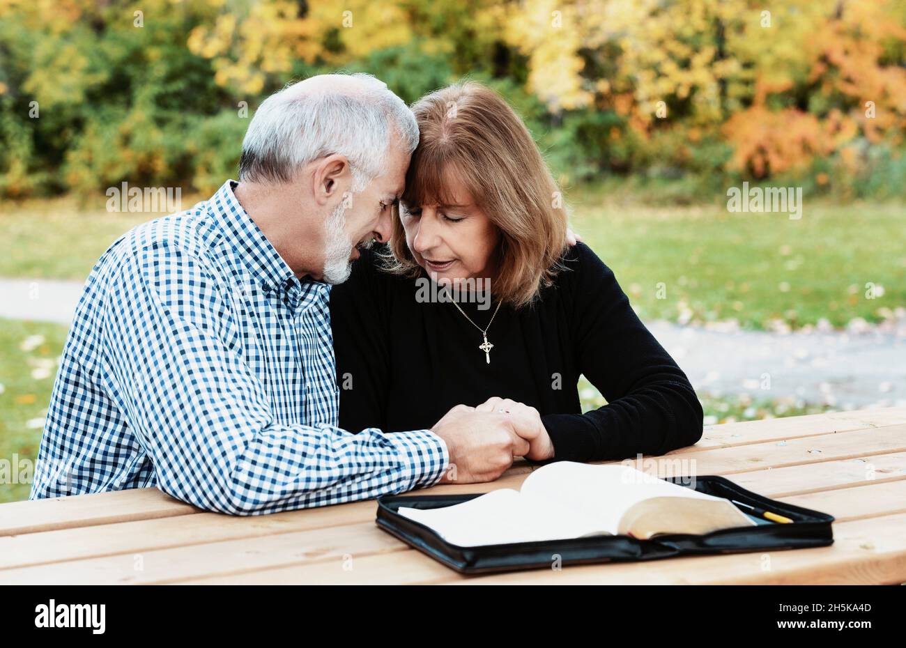 Ein reifes Paar, das sich an einem warmen Herbsttag in einem Stadtpark Zeit mit Hingabe teilt, indem es die Bibel studiert und gemeinsam an einem Picknicktisch betet Stockfoto