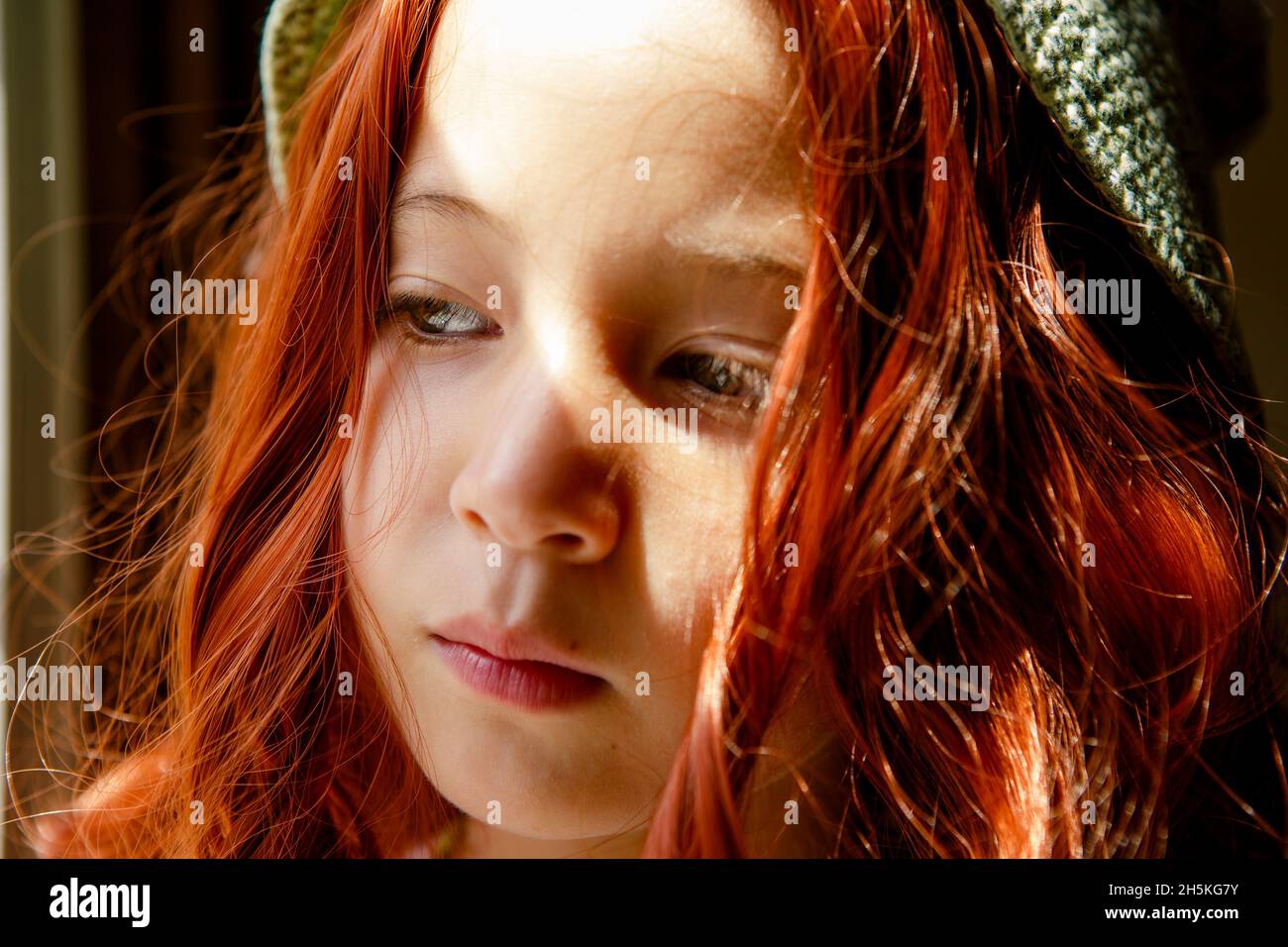 Ein ernsthaftes Kind mit leuchtend roten Haaren steht im Fensterlicht Stockfoto