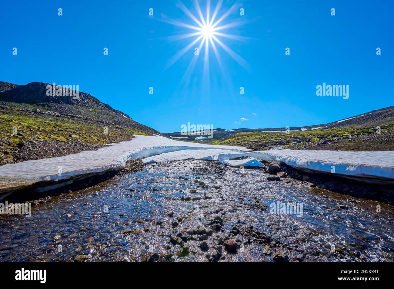 Bergbach mit Schneefeld und Sonneneinbruch in hellblauem Himmel im Sommer, Hellisheidi Eystri Bergpass in Nordisland Stockfoto