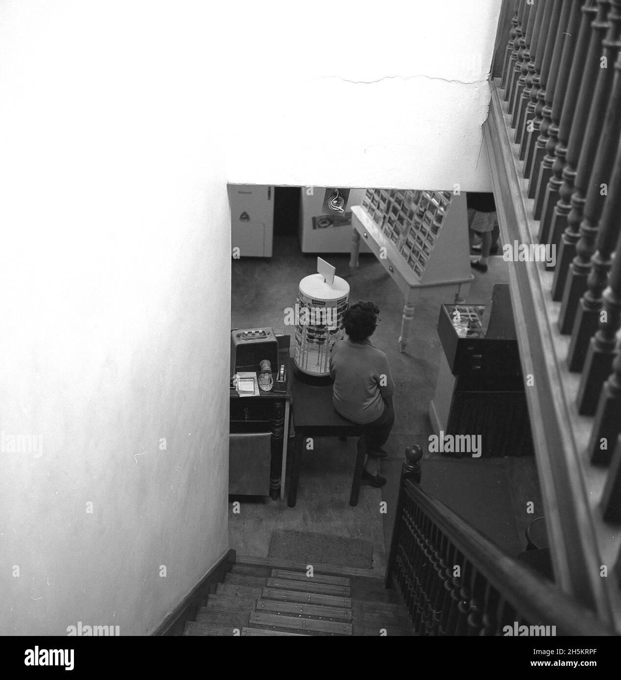 1960, historisch, von oben ein Blick über eine Treppe auf den Boden eines kleinen Elektrowarenladens, wo die Dame des Lagermeiters sitzt, auf einer Ecke eines Tisches, England, Großbritannien. Stockfoto
