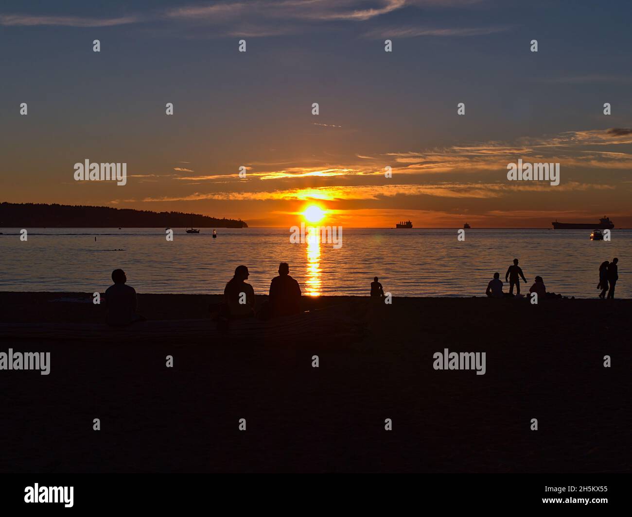 Atemberaubender Sonnenuntergang am beliebten Strand von English Bay in Vancouver, British Columbia, Kanada, mit Silhouetten von Menschen, die die Abendsonne und Boote genießen. Stockfoto