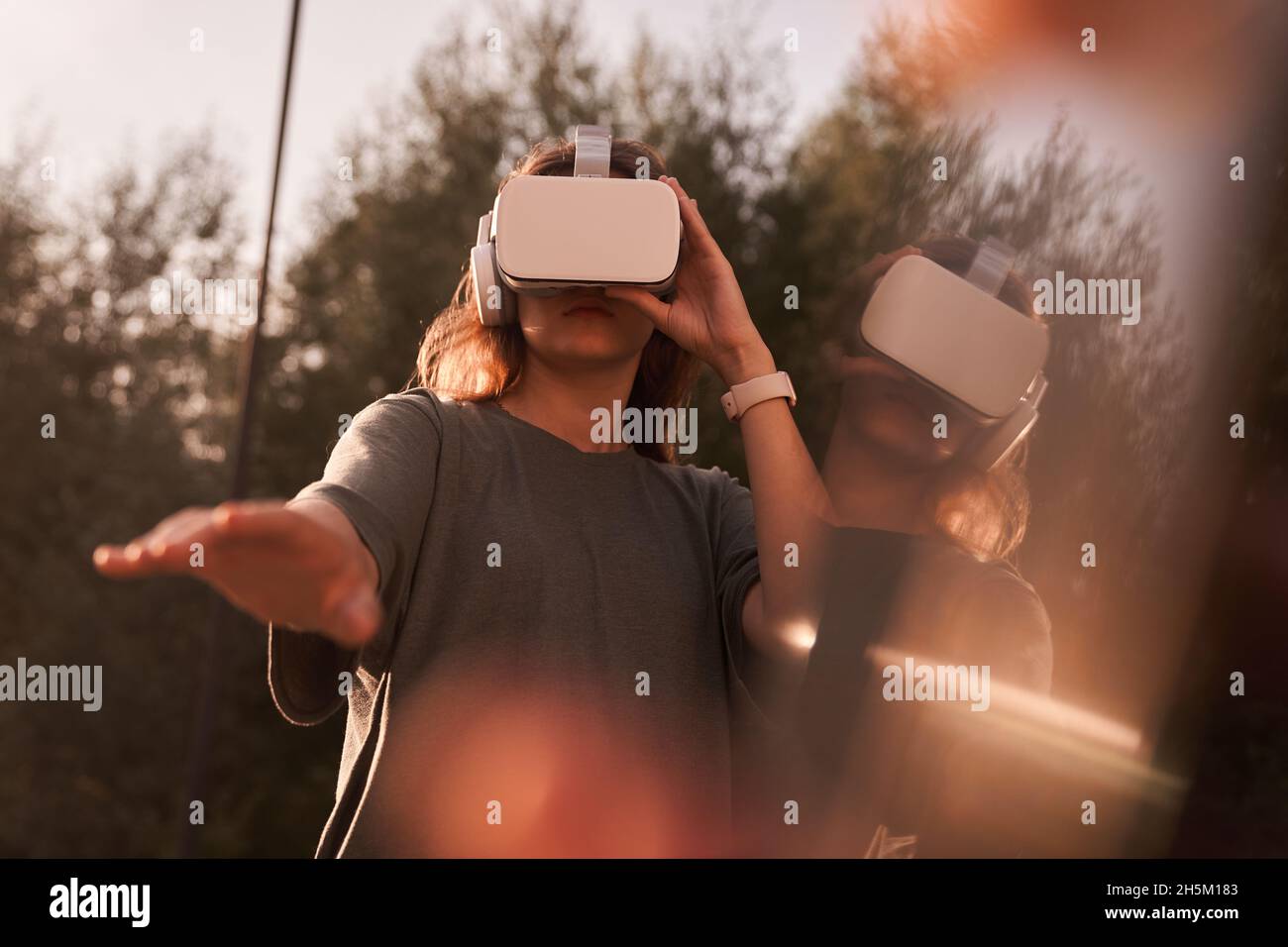 Nahaufnahme eines Mädchens, das mit den Händen eine Virtual-Reality-Brille auf dem Kopf hält und auf einem Hintergrund von grünen Bäumen steht. Er reicht eine Hand an die Kamera. Hochwertige Fotos Stockfoto