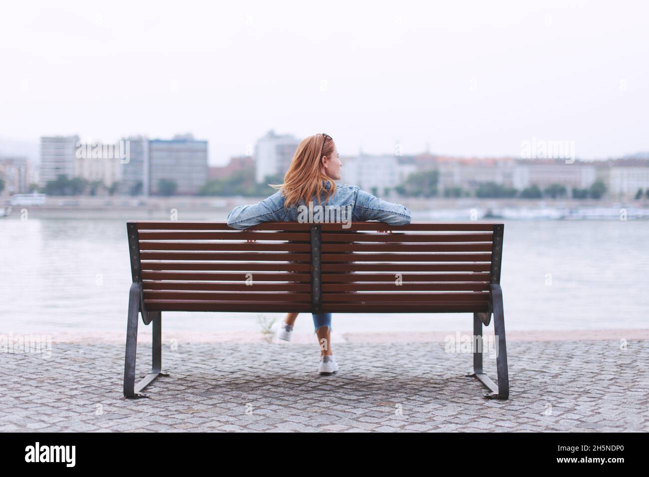 Junge Rothaarige Frau, die auf der Bank am Flussufer sitzt, Rückansicht Stockfoto
