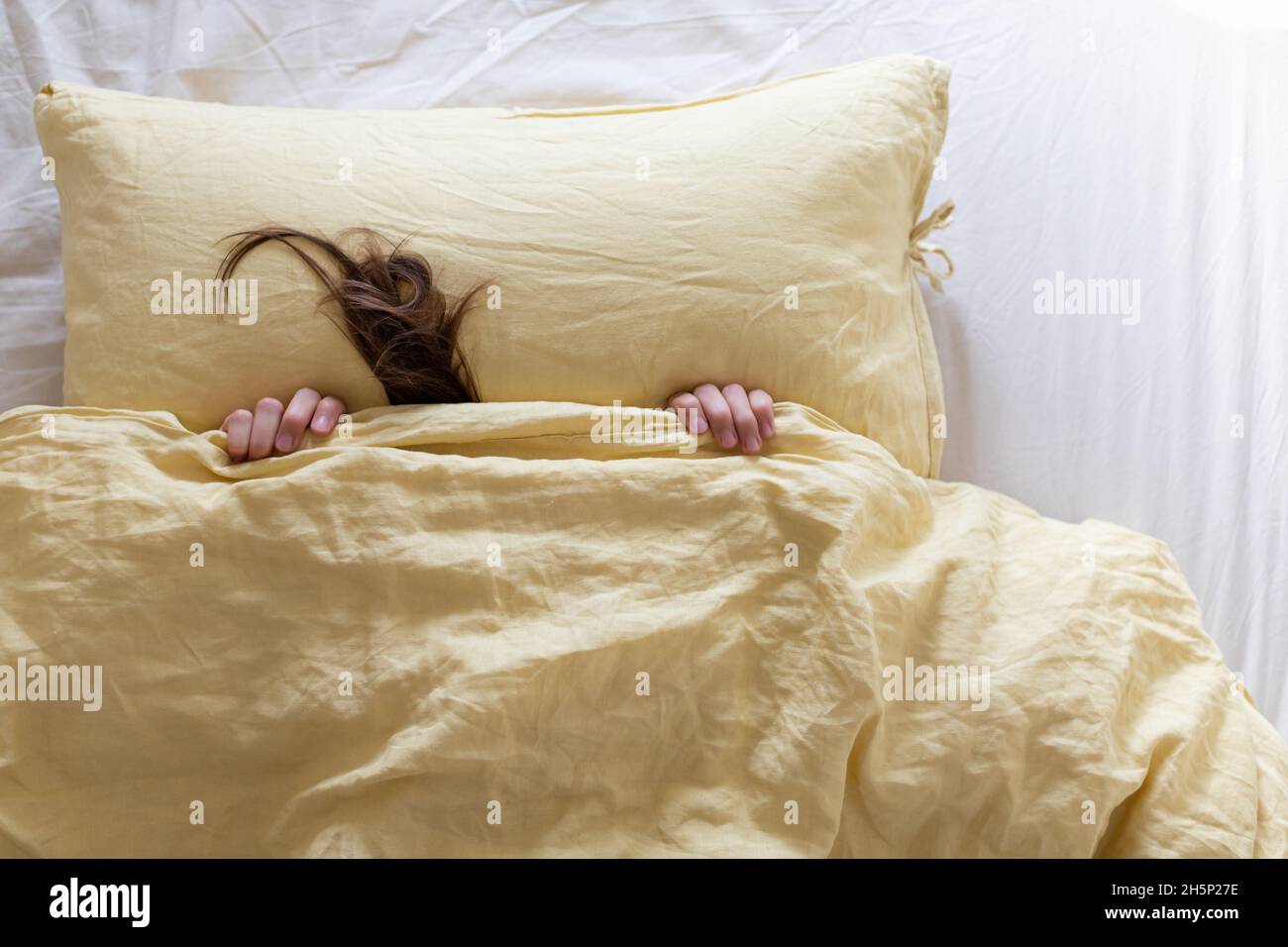 Frau im Bett ist mit einer gelben Decke mit ihrem Kopf bedeckt, man kann einen Teil ihrer Hände und Haare sehen. Das Konzept, sich vor Herbst und Winter zu verstecken. Stockfoto