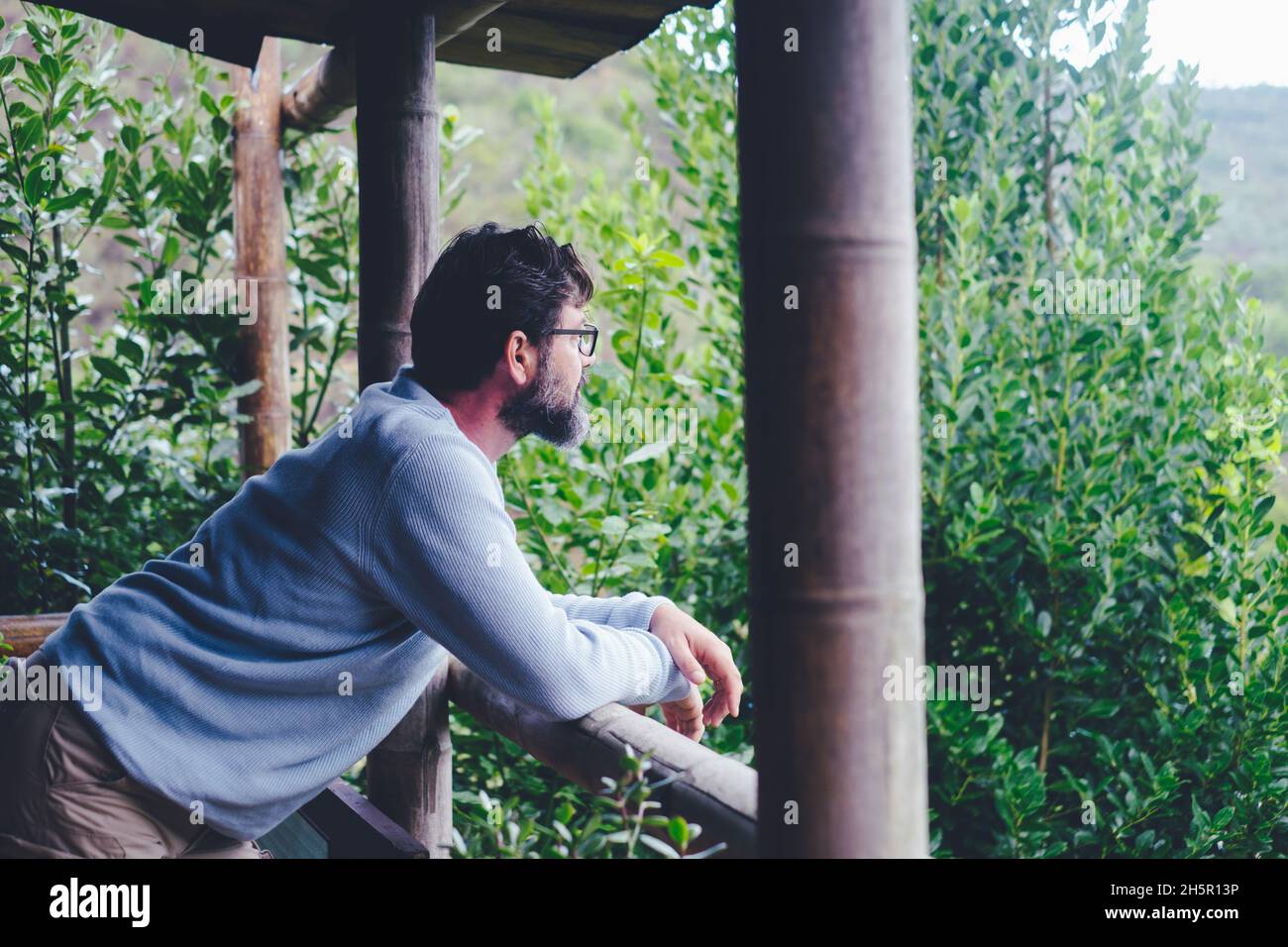 Ein Mann, der zu Hause auf der Terrasse stand und die grünen Bäume und Naturwälder draußen bewunderte. Entspannung und Lifestyle für Menschen. Erwachsener im Urlaub im Park Land Stockfoto Ein Mann, der zu Hause auf der Terrasse stand und die grünen Bäume und Naturwälder draußen bewunderte. Entspannung und Lifestyle für Menschen. Erwachsener im Urlaub im Park Land Stockfoto