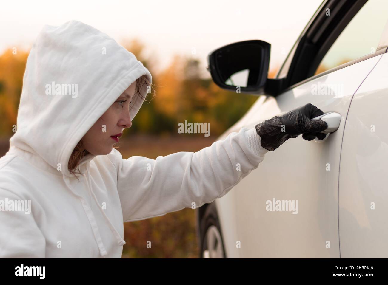 Eine schöne junge Frau in einem weißen Pullover mit Kapuze und schwarzen Handschuhen öffnet die Autotür, um sie zu stehlen. Selektiver Fokus. Nahaufnahme. Hochformat Stockfoto