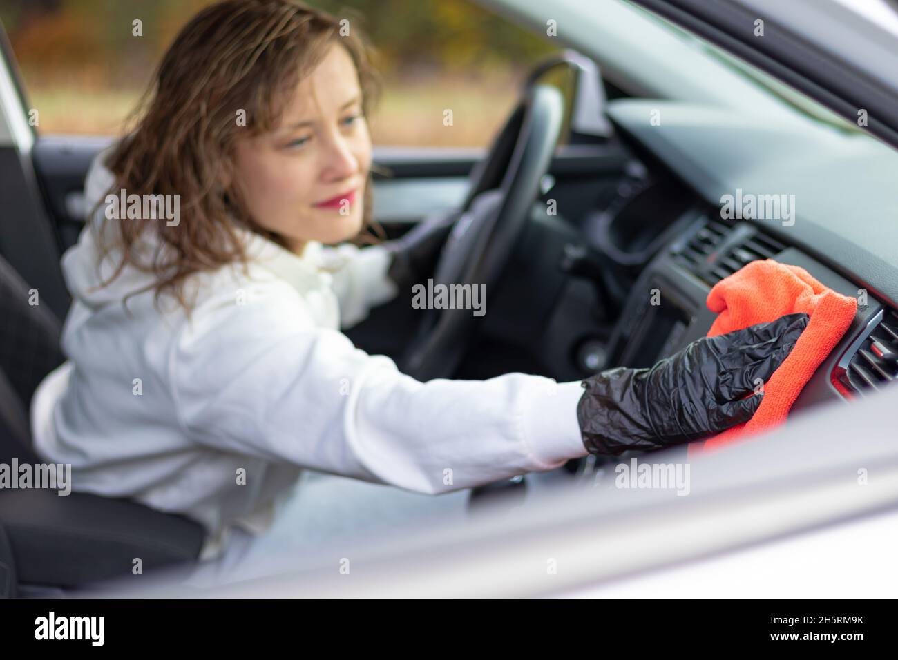 Eine schöne junge Autofahrerin in einer weißen Jacke und schwarzen Handschuhen wischt an einem strahlend warmen Herbsttag den Staub im Innenraum des Autos mit einem roten Lappen ab. Auswahl Stockfoto