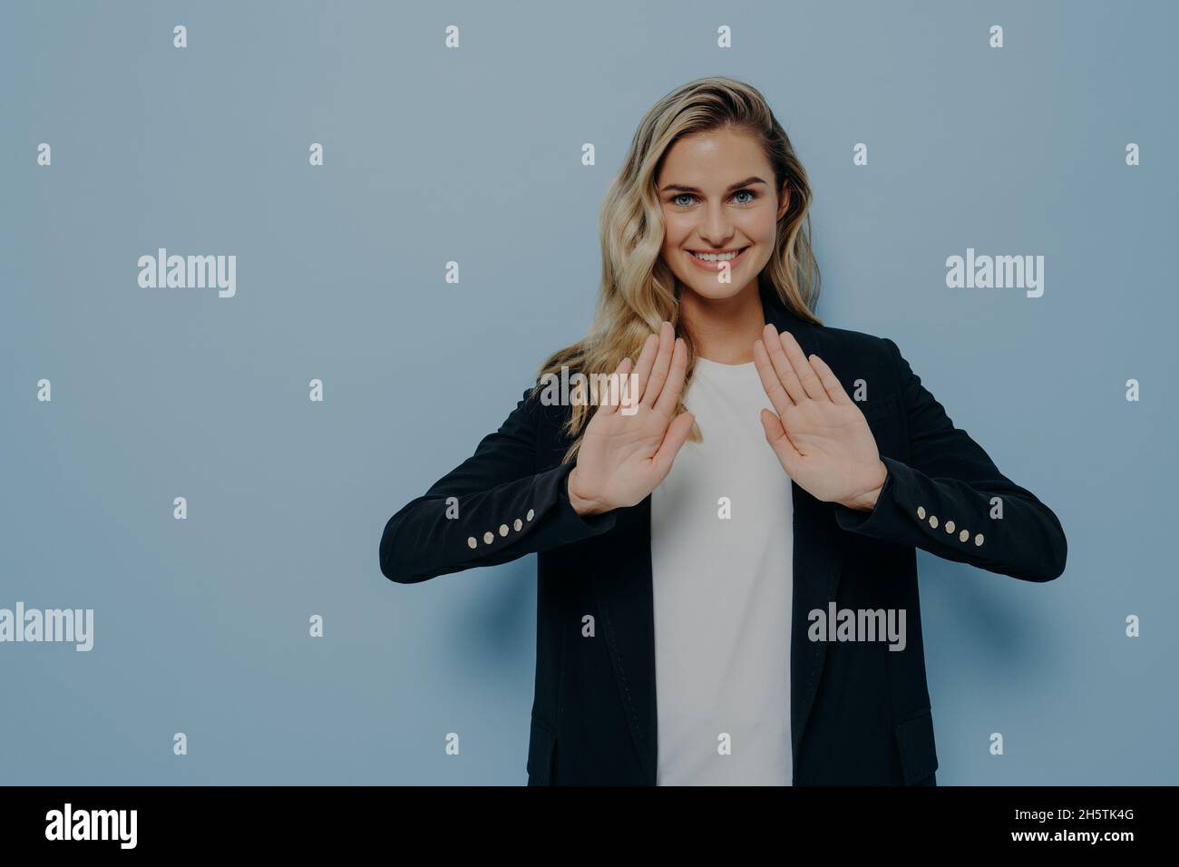 Lächelnde, höfliche junge Frau mit schwarzem Blazer über weißem T-Shirt, die die Hände in einer Stoppgeste hält Stockfoto