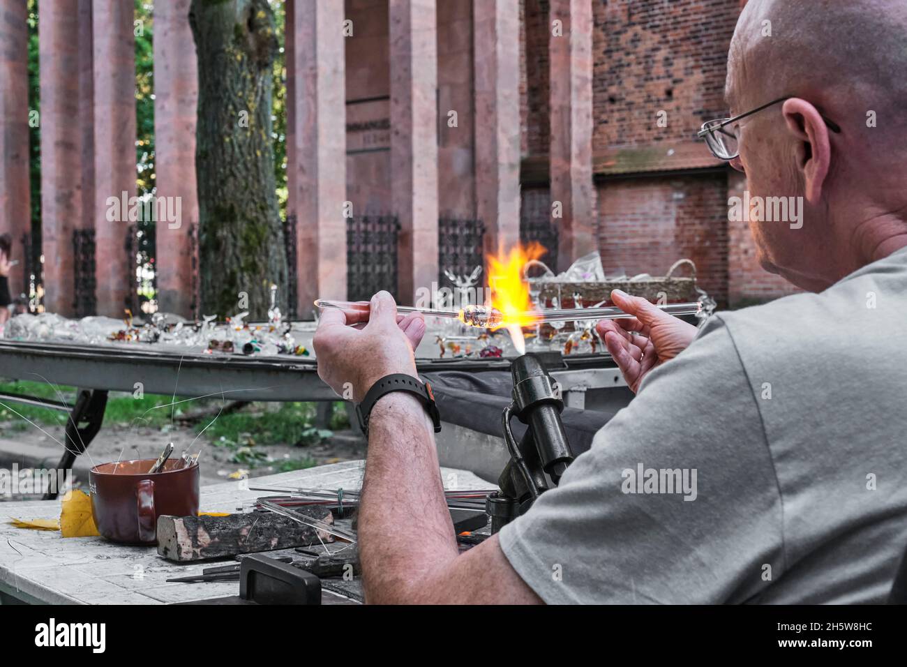 Hände eines reifen Glasbläsers, der Glasstab über der Brennerflamme hält. Prozess der künstlerischen Glasverarbeitung, Flammenarbeit. Stockfoto