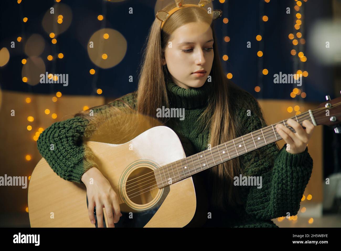 Ein schönes Teenager-Mädchen in weihnachtlichen Geweihen und ein grüner Pullover spielt Gitarre. Häusliche Umgebung, dunkler Hintergrund mit Bokeh aus Weihnachtslicht Stockfoto
