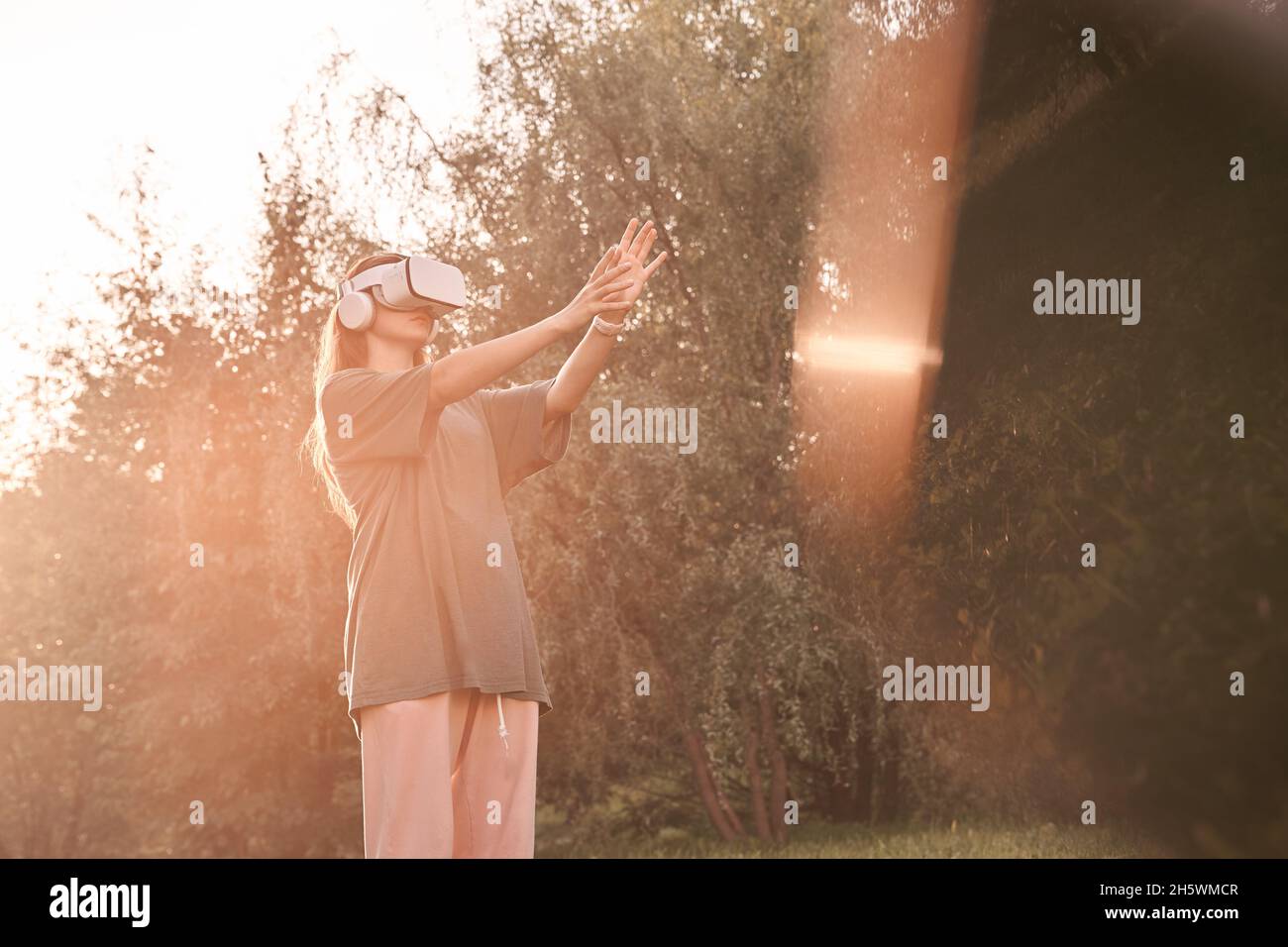 Ein Mädchen, das mit den Händen einen Virtual-Reality-Helm auf dem Kopf hält. Sie hält ihre Hände einem Sonnenstrahl entgegen. Hochwertige Fotos Stockfoto