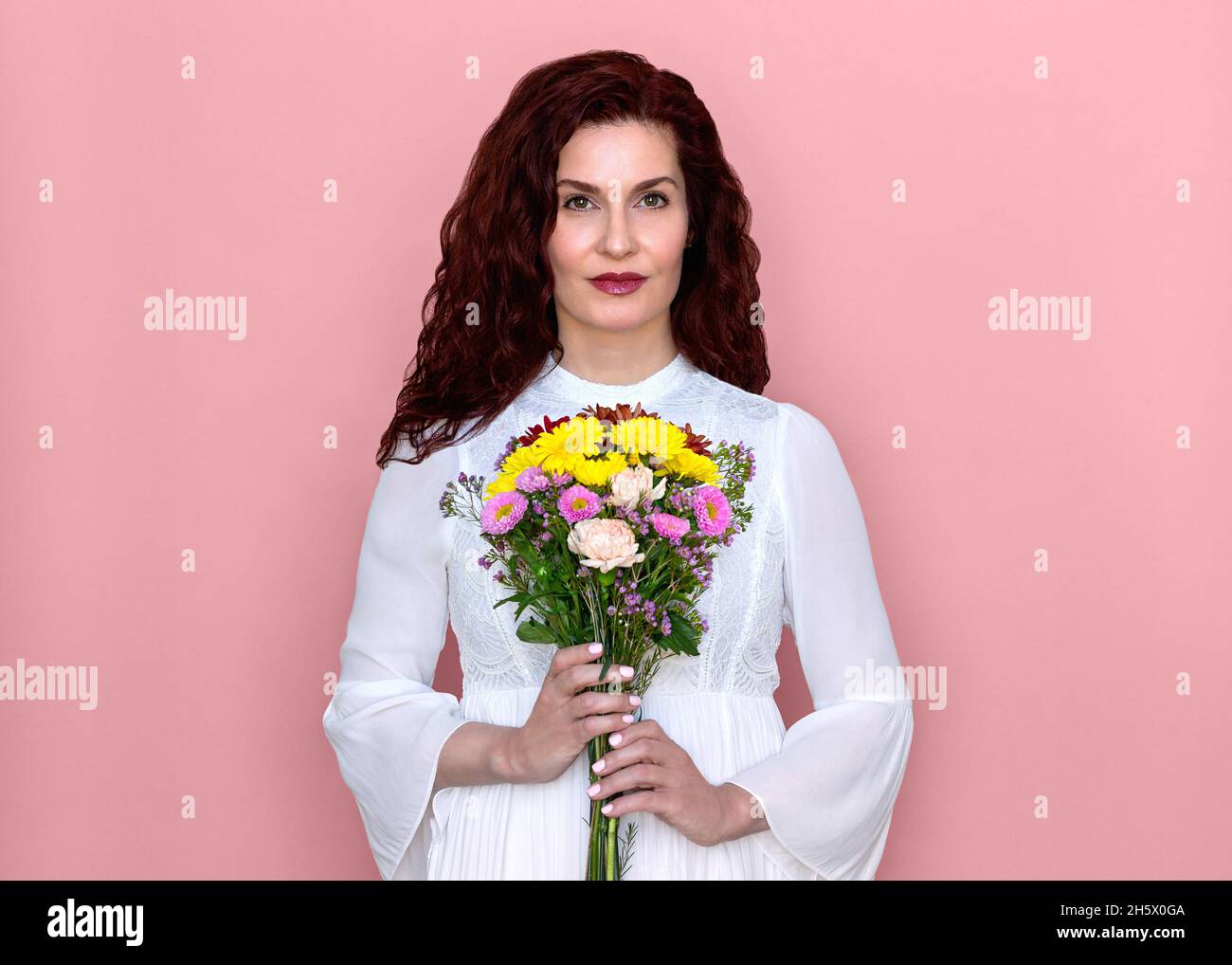 Frau hält Blumen vor ihrer Brust mit rosa Hintergrund. Romantische Studio Porträt der schönen Frau in weißem Kleid mit Blumenstrauß. Stockfoto