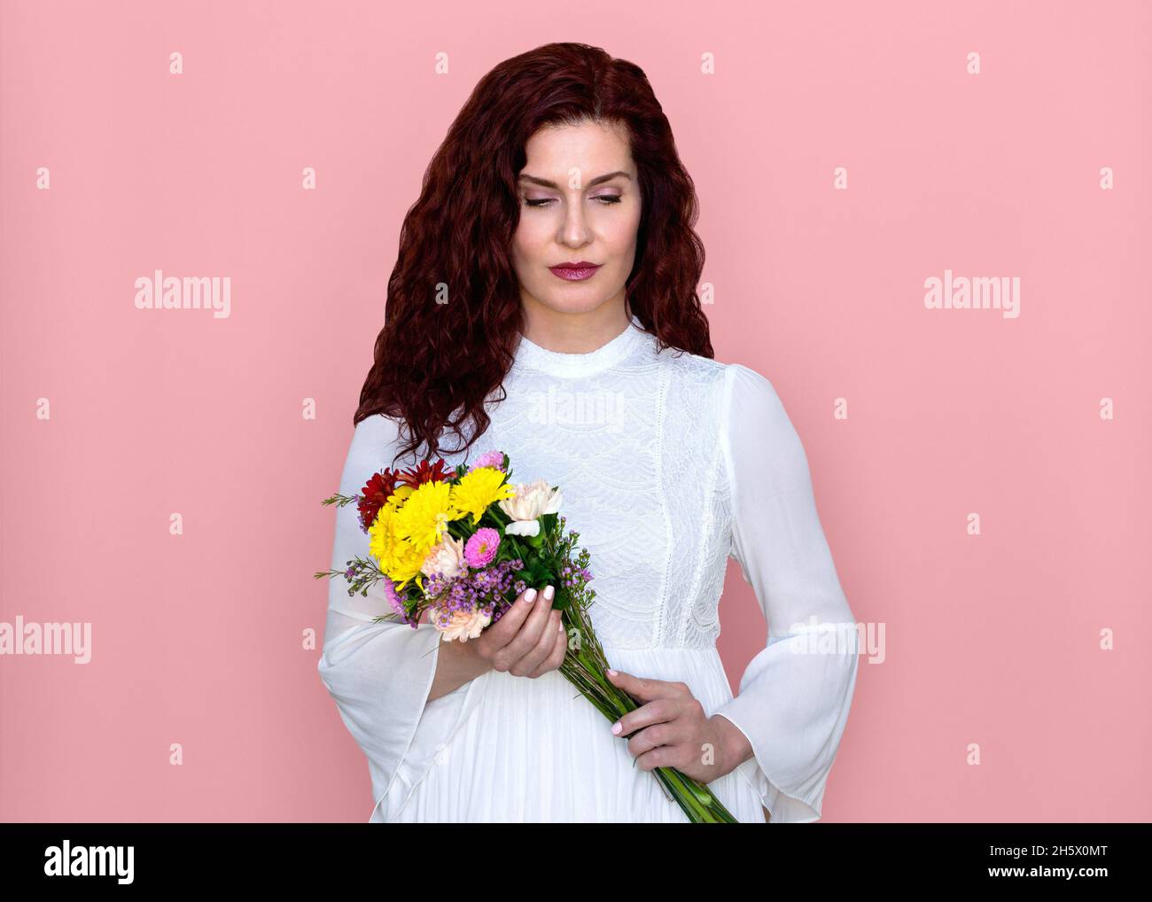 Frau, die nach unten schaut, hält Blumenstrauß mit rosa Hintergrund. Schöne nachdenkliche Frau in romantischen weißen Kleid mit Blumenstrauß. Stockfoto