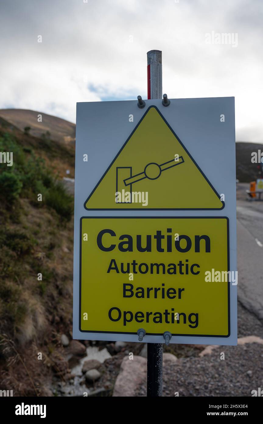 Gelbes und weißes Schild mit der Aufschrift „Achtung Automatische Barriere in Betrieb“. Barriere-Symbol. Verschwommener Hintergrund von Barriere, Straße und Bergen. Stockfoto