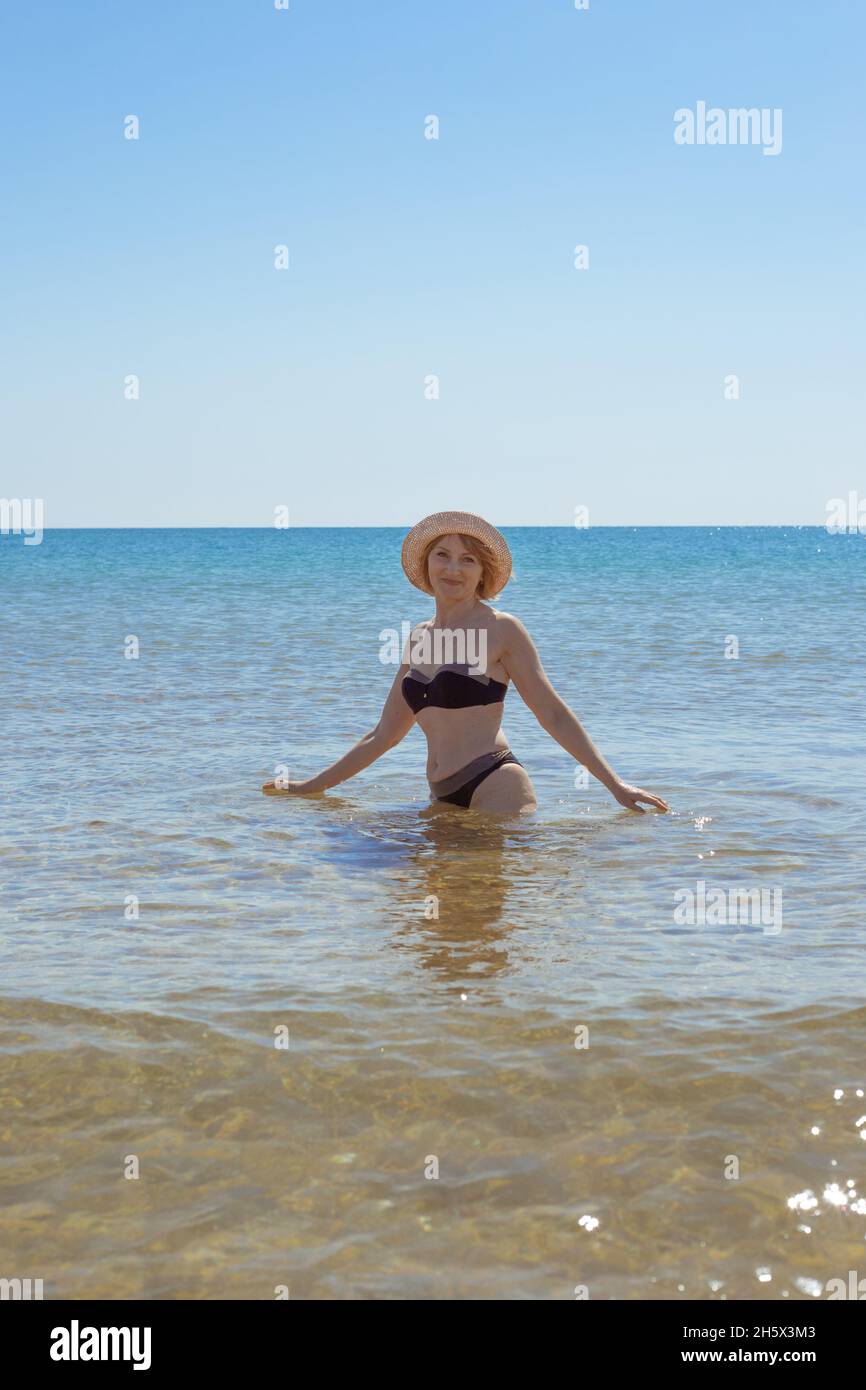 Europäische Frau im Badeanzug und hüftlangen Hut im Meer lächelt und schaut in die Kamera. Urlaub am Meer. Stockfoto