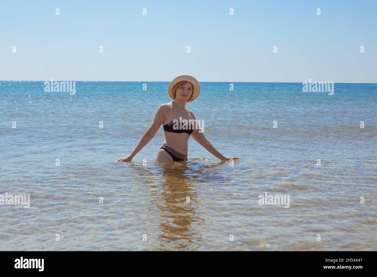 Europäische Frau im Badeanzug und hüftlangen Hut im Meer lächelt und schaut in die Kamera. Urlaub am Meer. Stockfoto