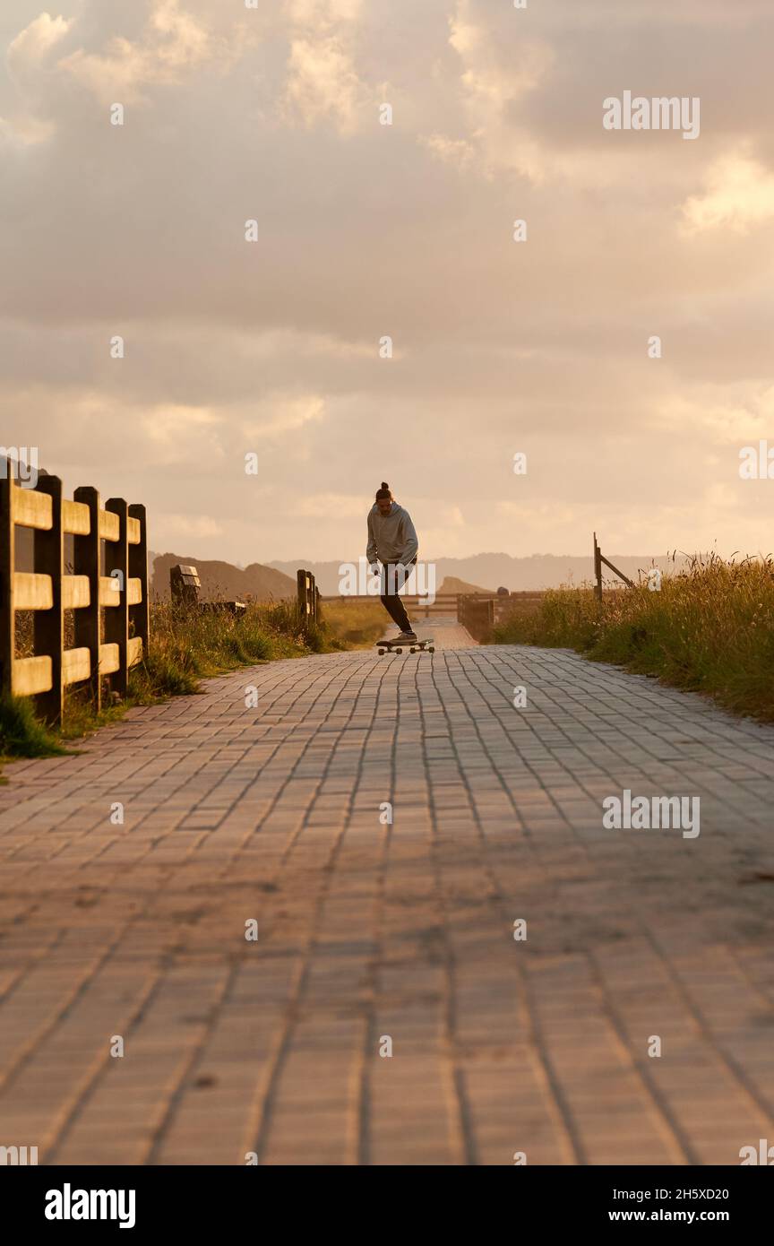 Ganzer Körper von aktiven männlichen Reiten Longboard entlang gepflasterten langen Weg unter bewölktem Himmel bei Sonnenuntergang am Abend Stockfoto