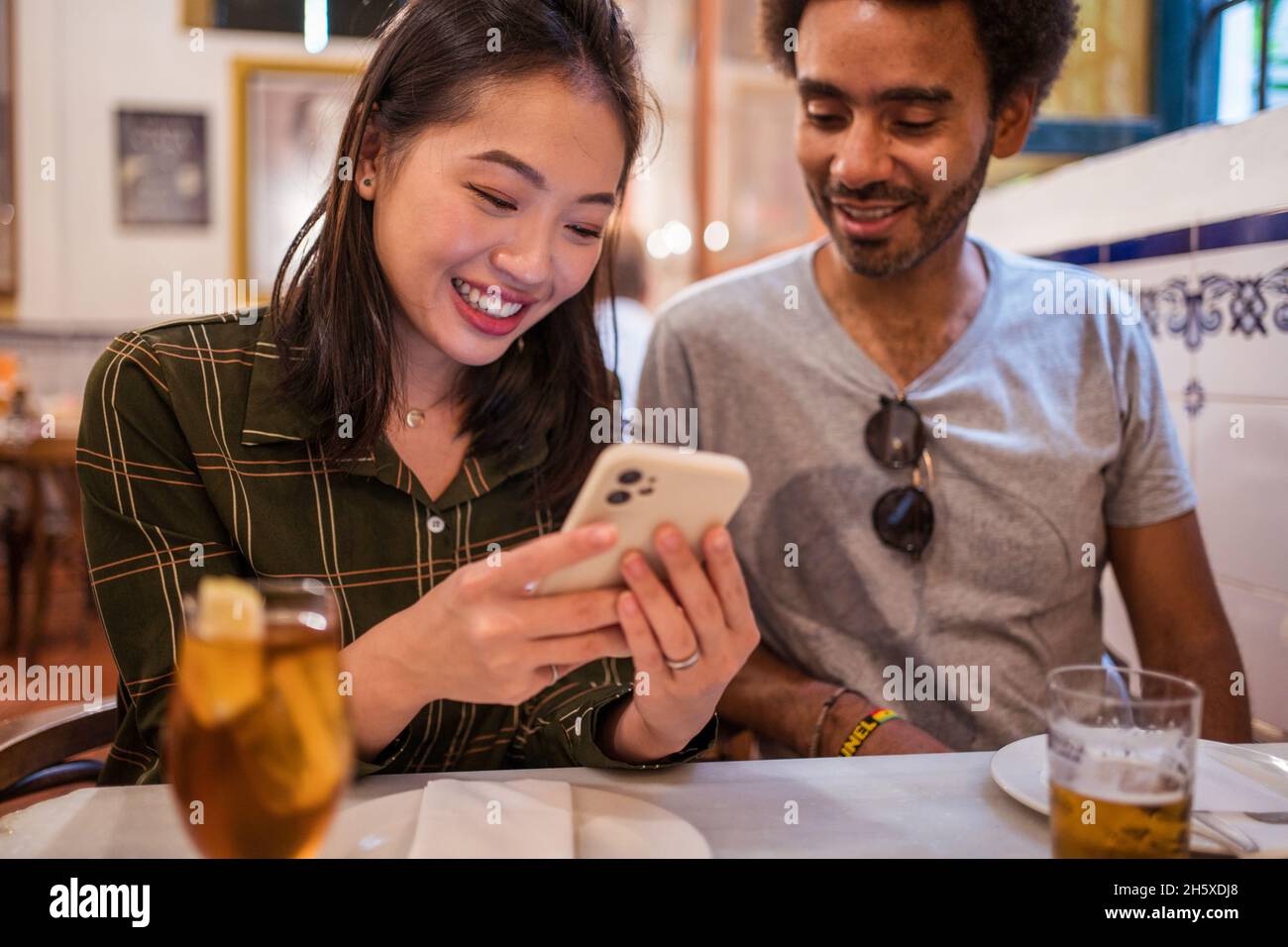 Junge asiatische Frau, die Fotos auf dem Smartphone einem ethnischen Freund zeigt, der am Tisch im Restaurant sitzt Stockfoto