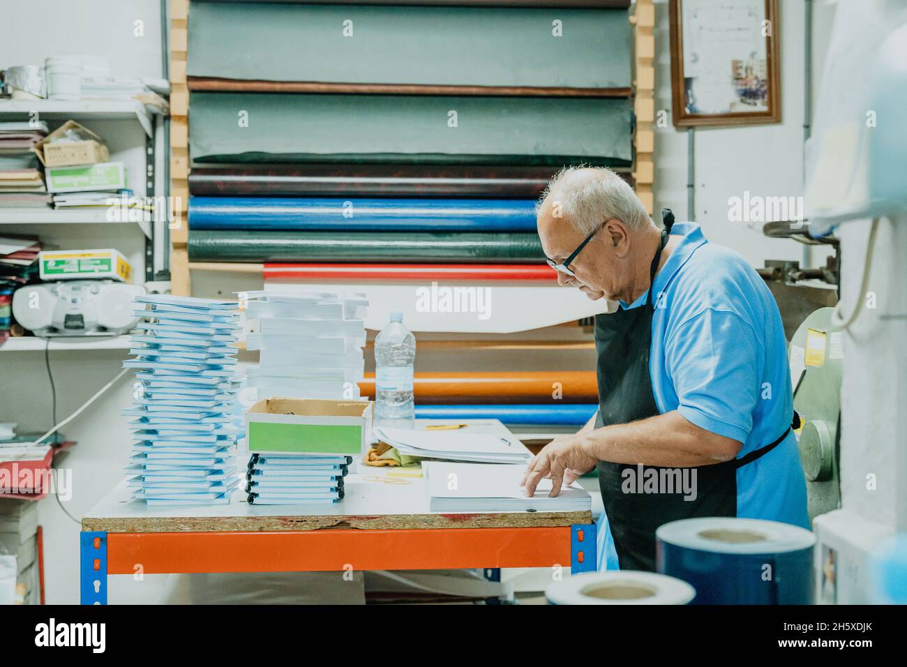 Seitenansicht des konzentrierten älteren männlichen Handwerkers mit weißen Haaren in Schürze und Brillen, die am Tisch mit Papierstapel und Notizbuch im Druck stehen Stockfoto