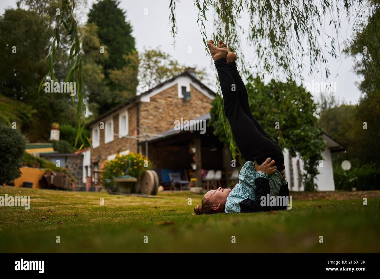 Ganzkörper-Seitenansicht des barfuß-Mädchens, das Ardha Sarvangasana auf einem Rasen im Hinterhof in der Nähe von Gebäuden auf dem Land anführt Stockfoto