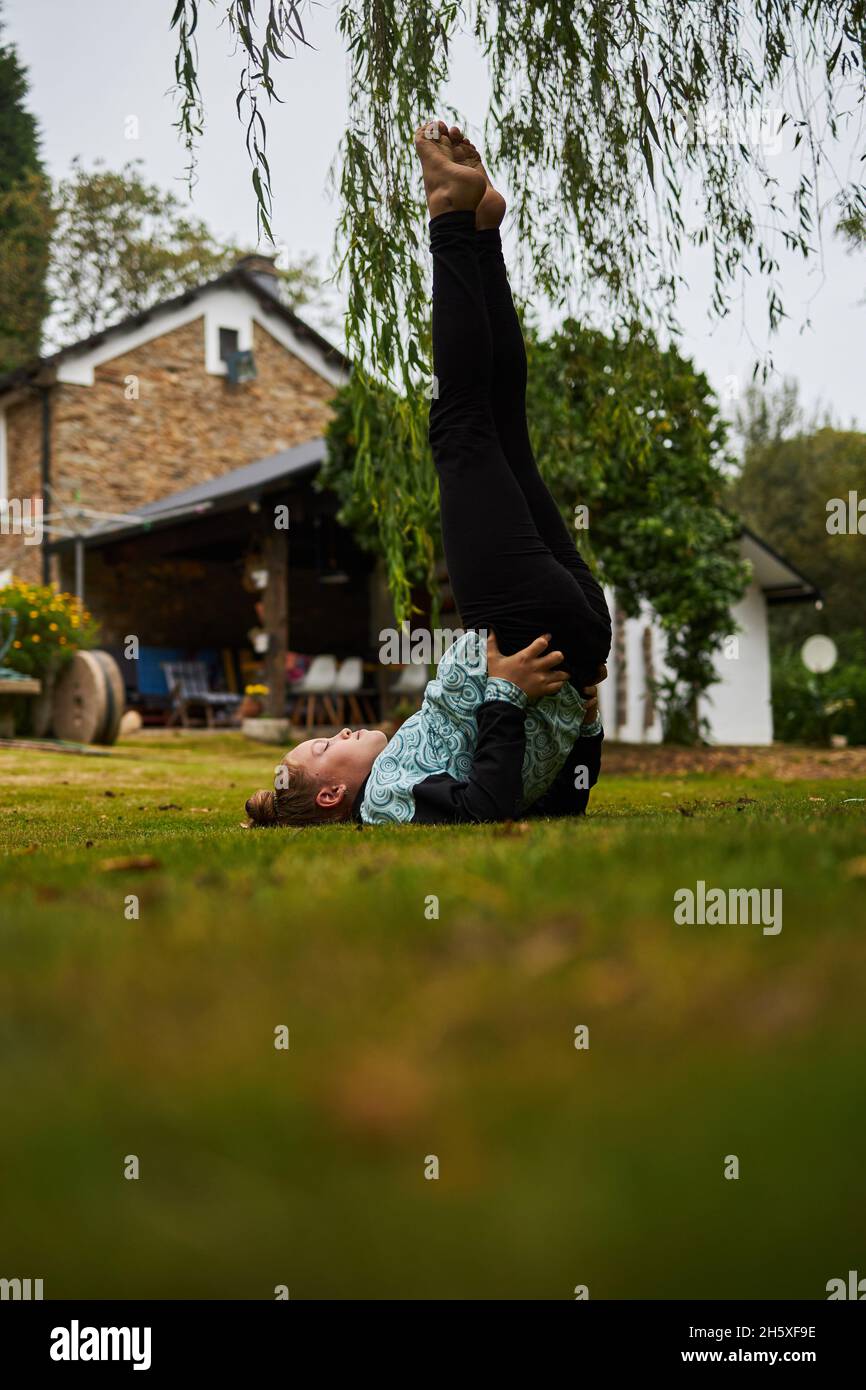 Ganzkörper-Seitenansicht des barfuß-Mädchens, das Ardha Sarvangasana auf einem Rasen im Hinterhof in der Nähe von Gebäuden auf dem Land anführt Stockfoto