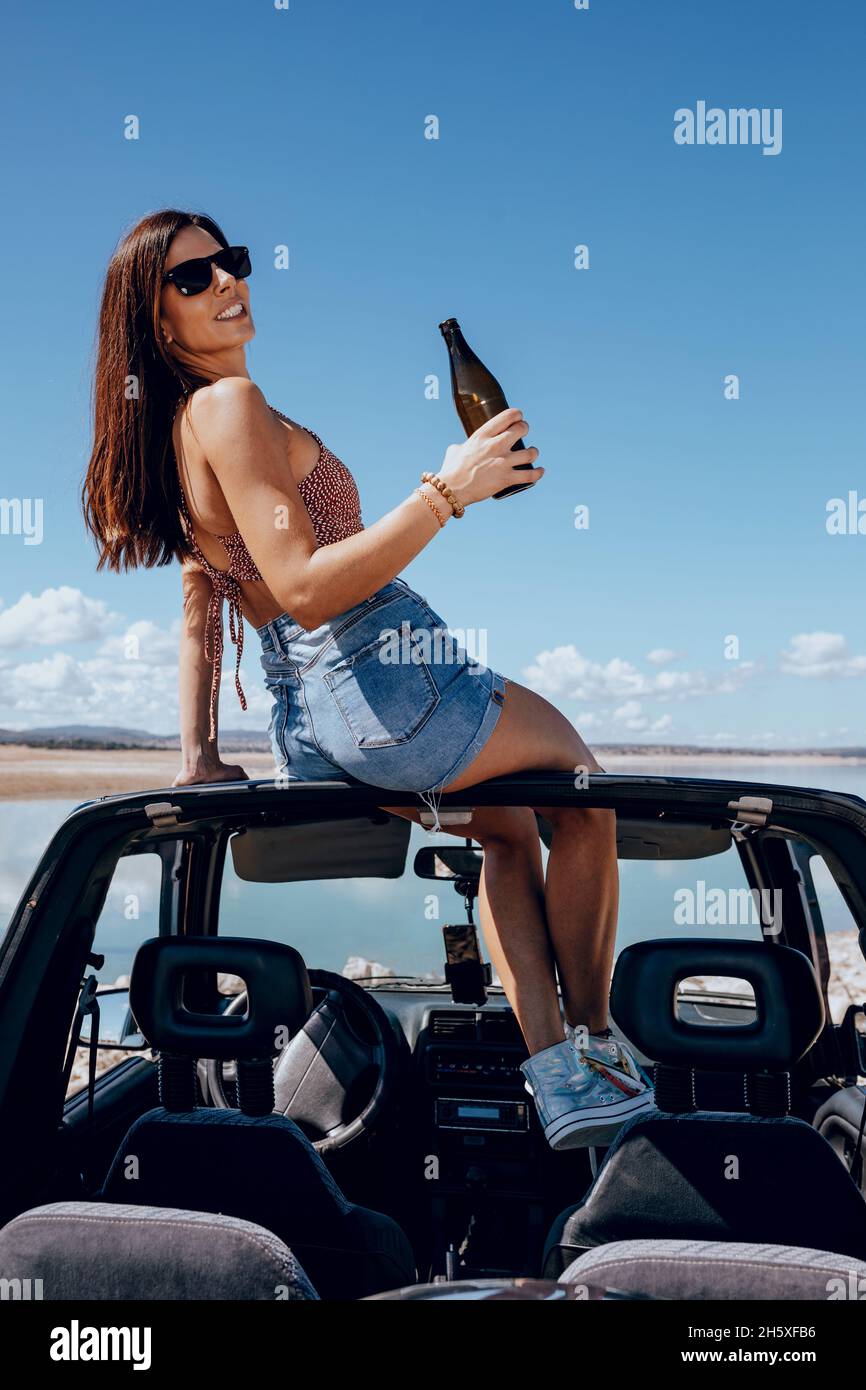 Ganzer Körper von glücklichen jungen Frauen im Sommer-Outfit und Sonnenbrille, die Bier hält, während sie auf dem Dach eines Safarikarons am Ufer des Flusses sitzen Stockfoto