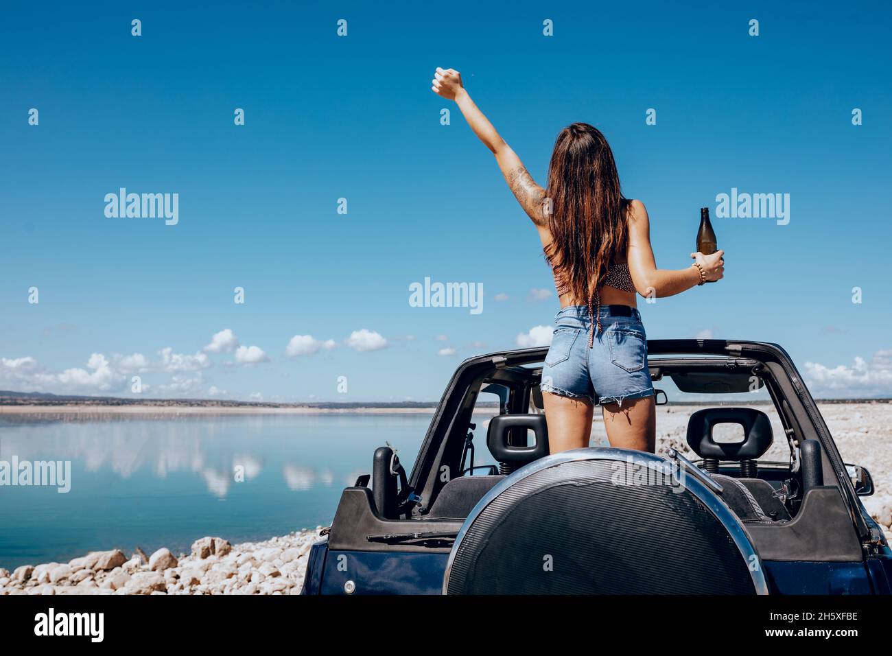 Rückansicht einer aufgeregten Frau im Sommeroutfit, die die Hand des Bieres hebt, während sie auf dem Dach eines Safarikarufers am Ufer des Flusses steht Stockfoto