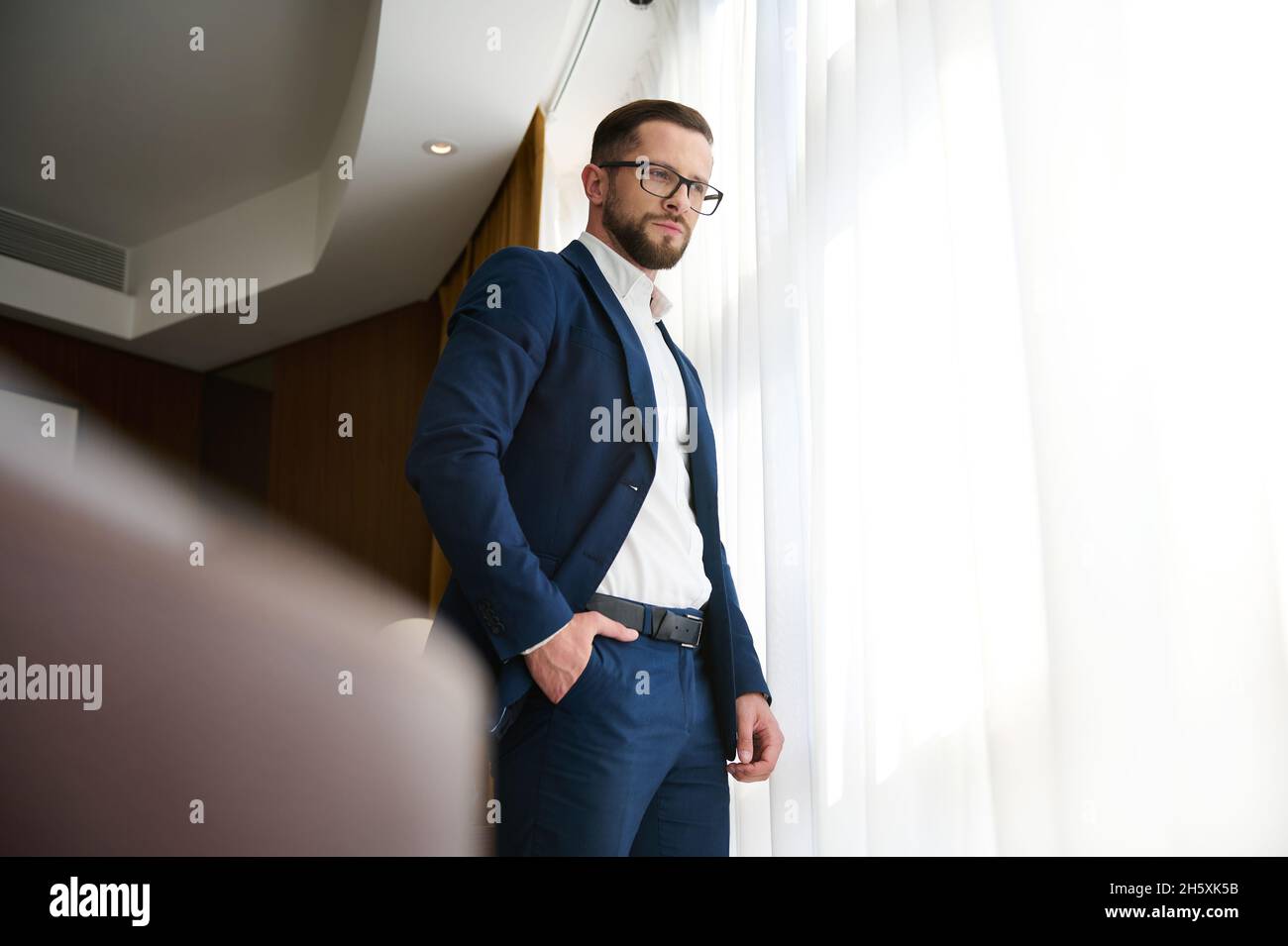 Gutaussehender wohlhabender Geschäftsmann, erfolgreicher Business-Investor, Unternehmer in Business-Anzug, steht mit Händen in der Tasche Blick auf das Stadtbild durch Stockfoto