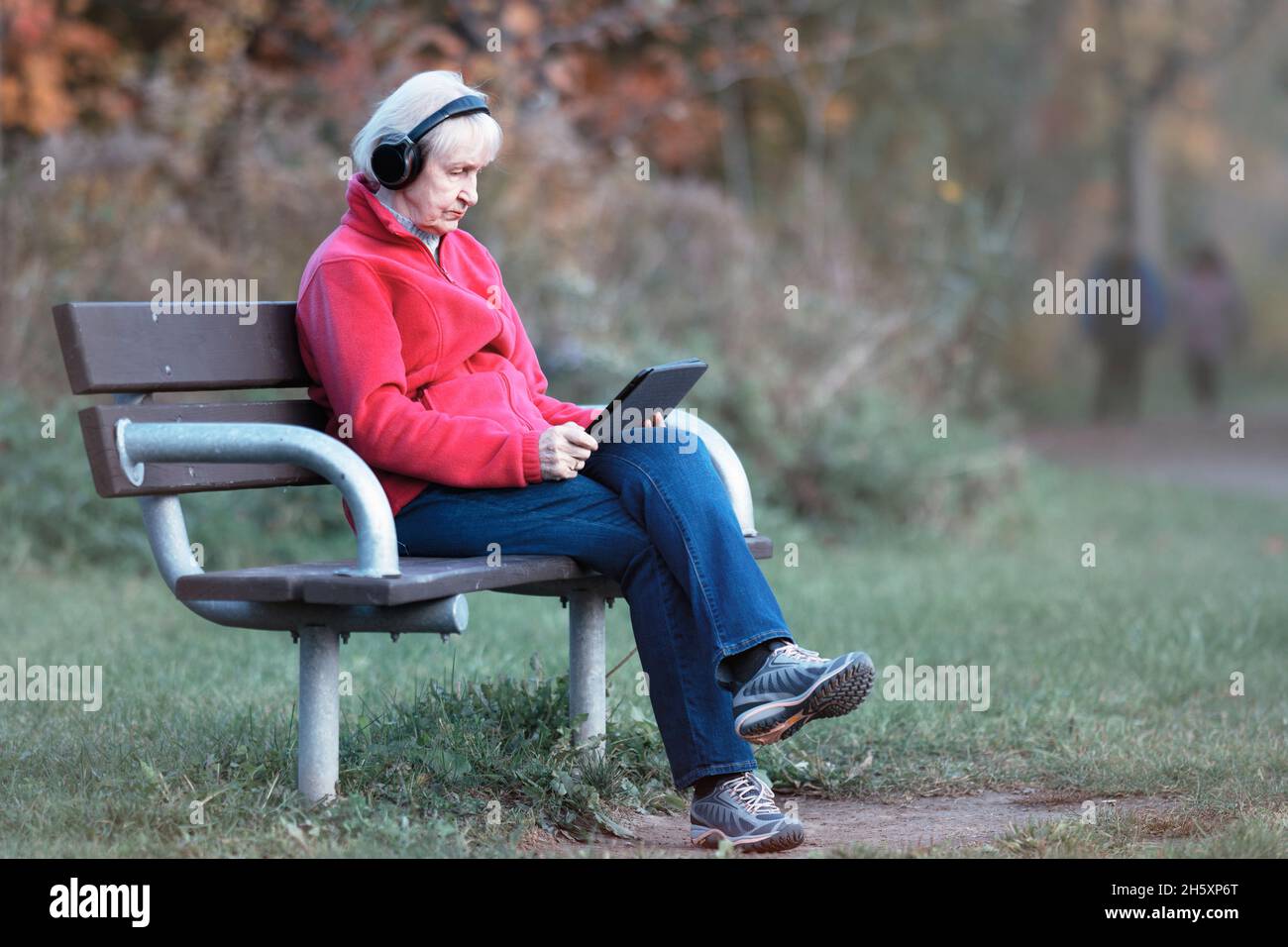 Ältere ältere kaukasische Frau, die allein auf der Parkbank sitzt und Kopfhörer trägt und Tablet hält Stockfoto