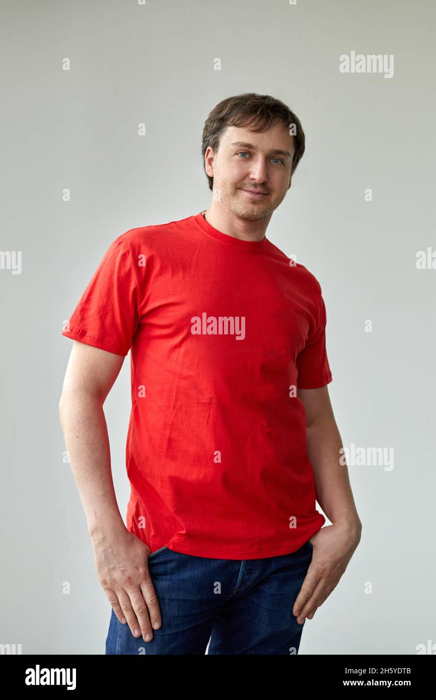 Positive junge männliche Modell in lässigem roten T-Shirt und Jeans Blick auf die Kamera während des Stehens. Junger Mann im roten T-Shirt auf weißem Hintergrund Stockfoto