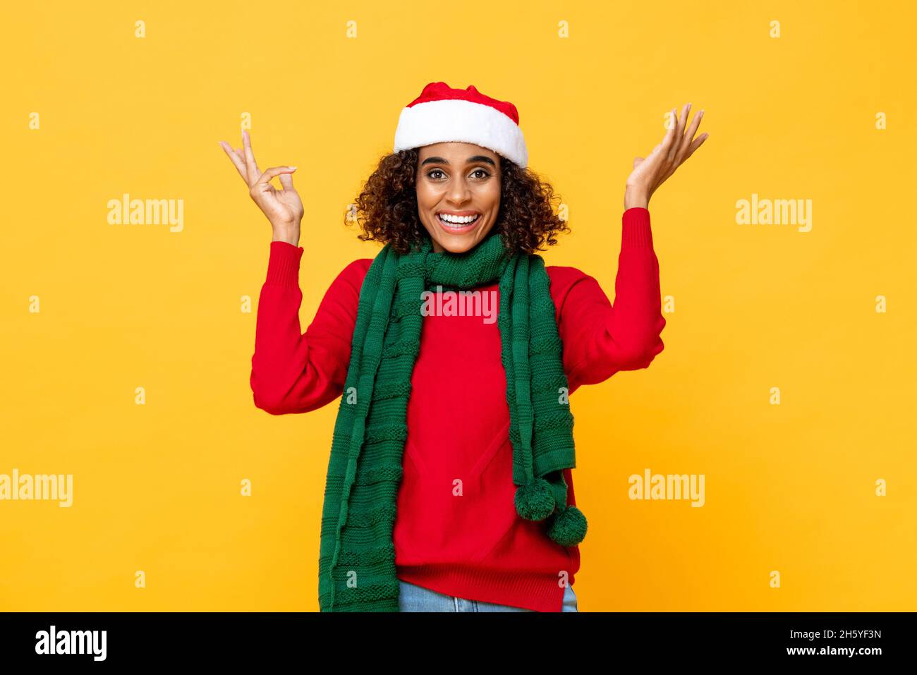 Überraschte Frau in Weihnachtskleidung lächelnd und die Hände hochhebend in gelbem Studio isolierten Hintergrund Stockfoto