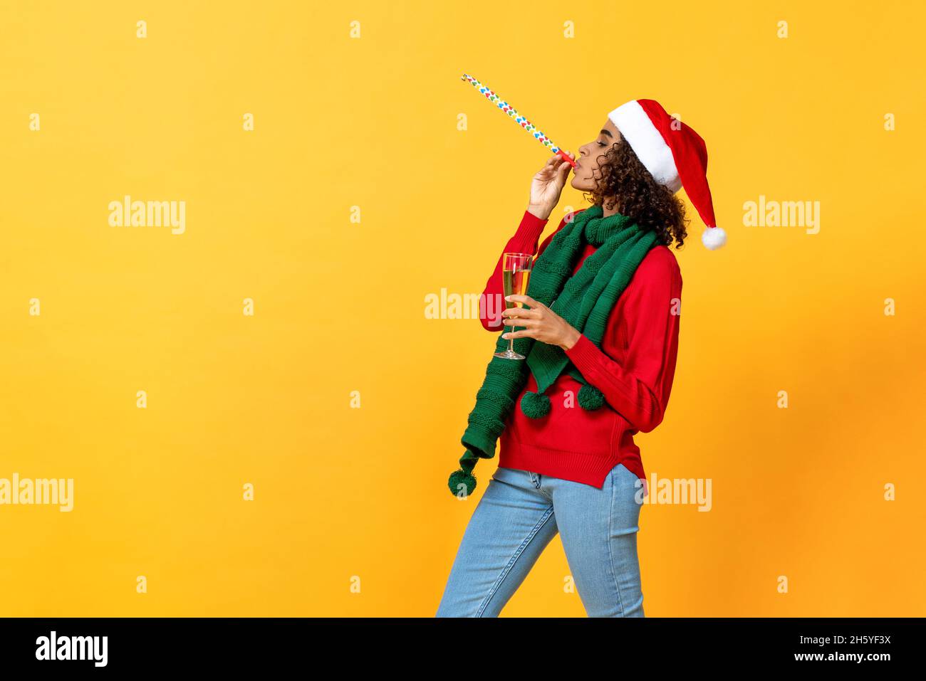 Happy African American Frau hält Champagner Glas und weht Party Horn zelebrierten Weihnachten in gelb isoliert Studio Hintergrund Stockfoto
