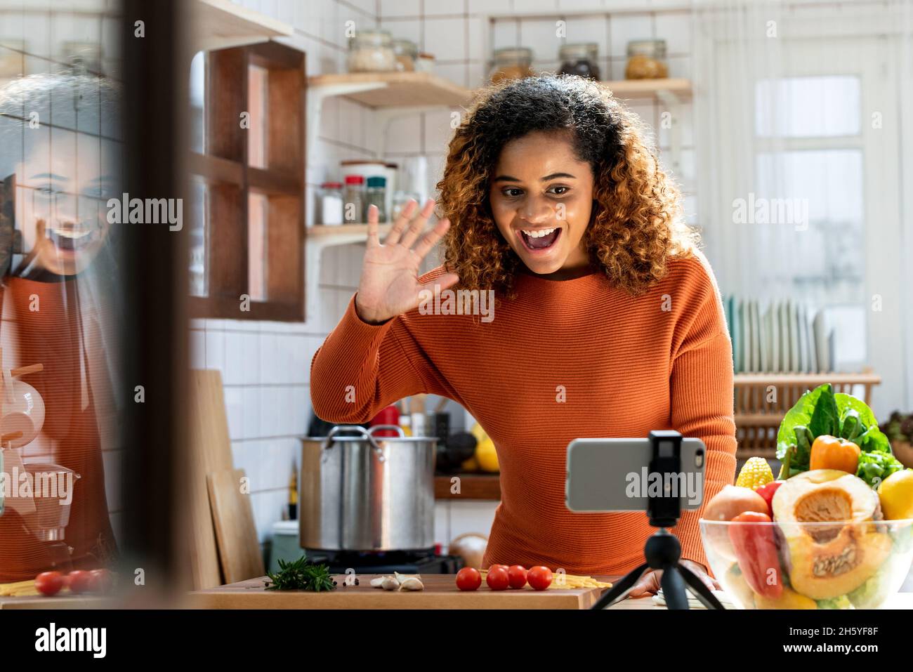 Junge afroamerikanische Frau, die auf das Mobiltelefon schaut und in den sozialen Medien in der Küche zu Hause Hallo sagt, virtuelles Kochkurs-Konzept Stockfoto