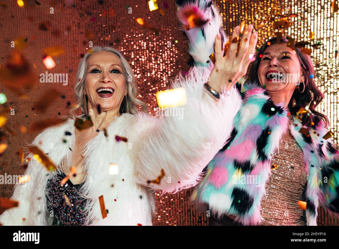 Zwei fröhliche ältere Frauen in Pelzmänteln, die unter Konfetti vor glitzerndem Hintergrund tanzen Stockfoto