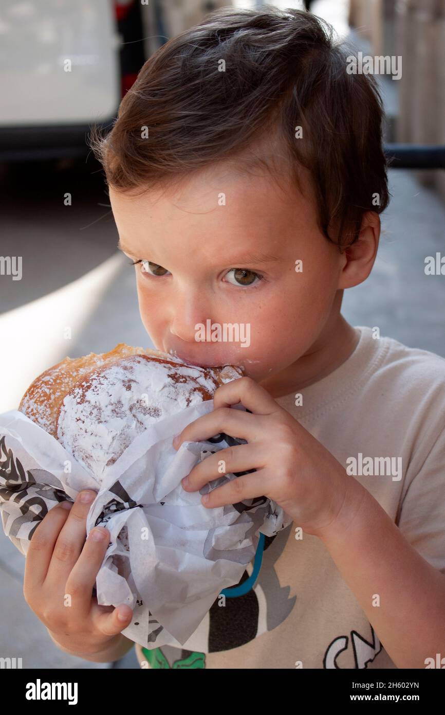 Das Kind isst eine große runde Brötchen. Porträt des Kindes, das ein Brioche-Brötchen mit Puderzucker beißt Stockfoto