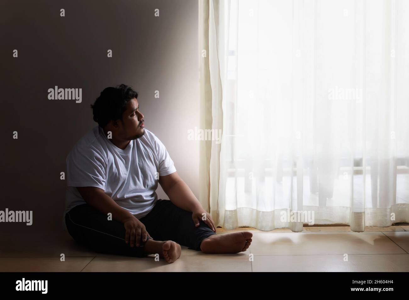 Ein fetter Mann sitzt traurig und schaut auf die Vorhänge in seinem Zimmer. Stockfoto