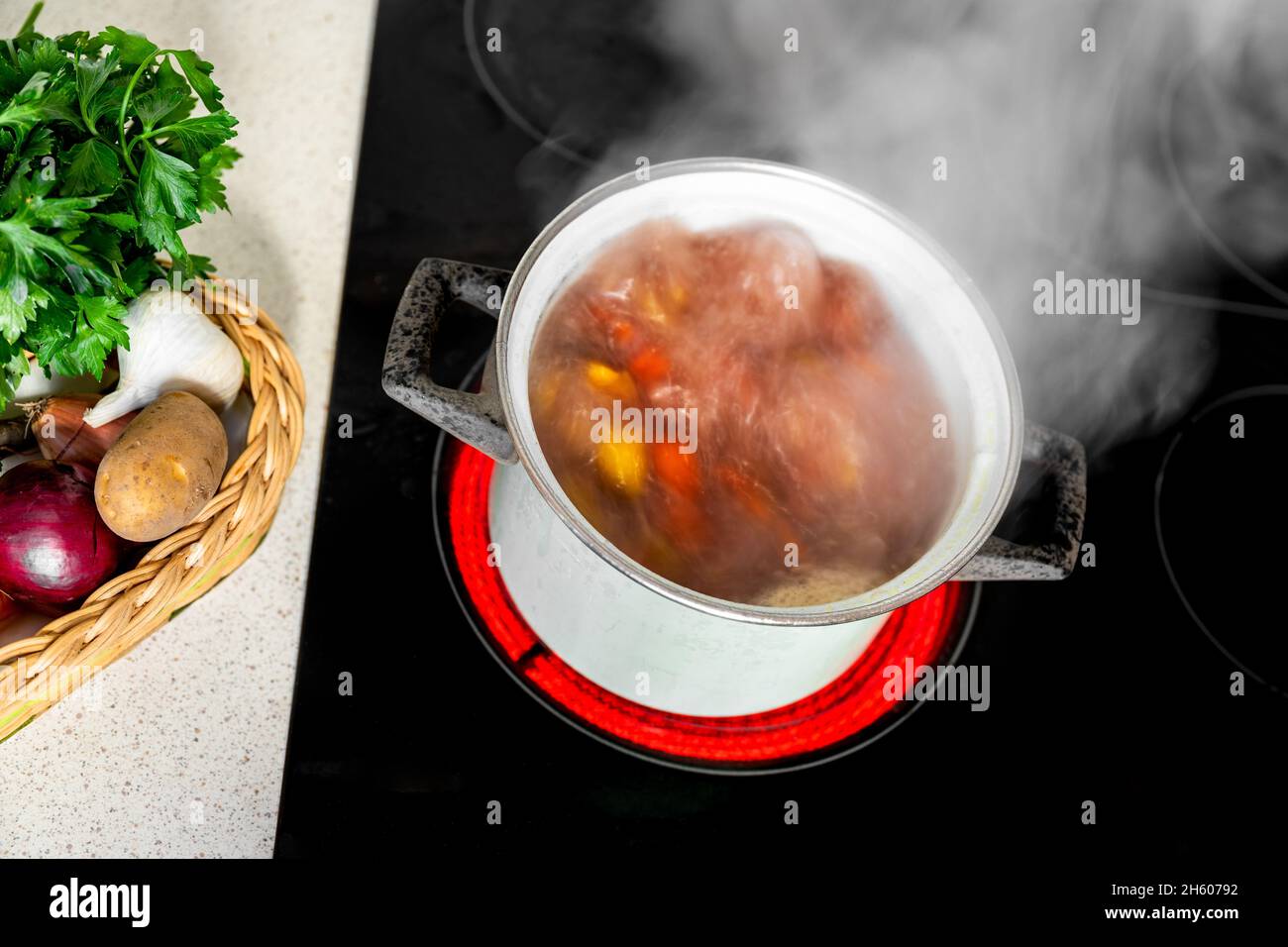 Karottensuppe kochen. Kochende Brühe mit bunten Karotten, Zwiebeln und Kartoffeln im Topf. Suppe sprudelt und raucht auf heißem Keramikkochfeld, frisches Gemüse. Stockfoto