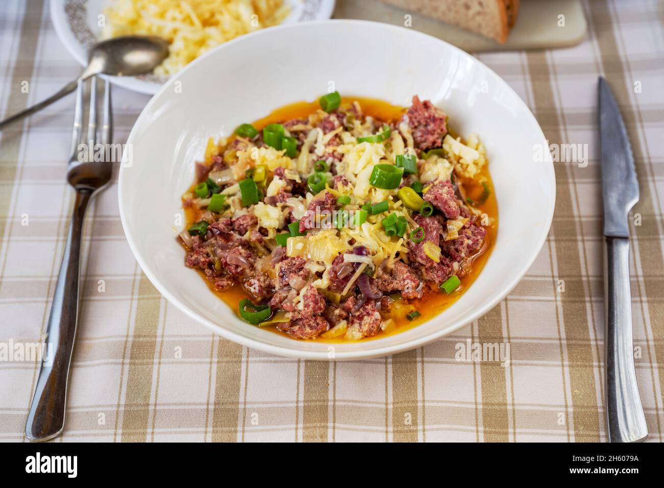 Hackfleisch aus Rindfleisch mit Zwiebeln und Lauch, geriebenem Käse, Besteck auf Tischdecke. Traditionelles Schweizer Gericht „Benediktiener eintopf“. Reichhaltiges Essen. 1-Topf-Mahlzeit. Stockfoto