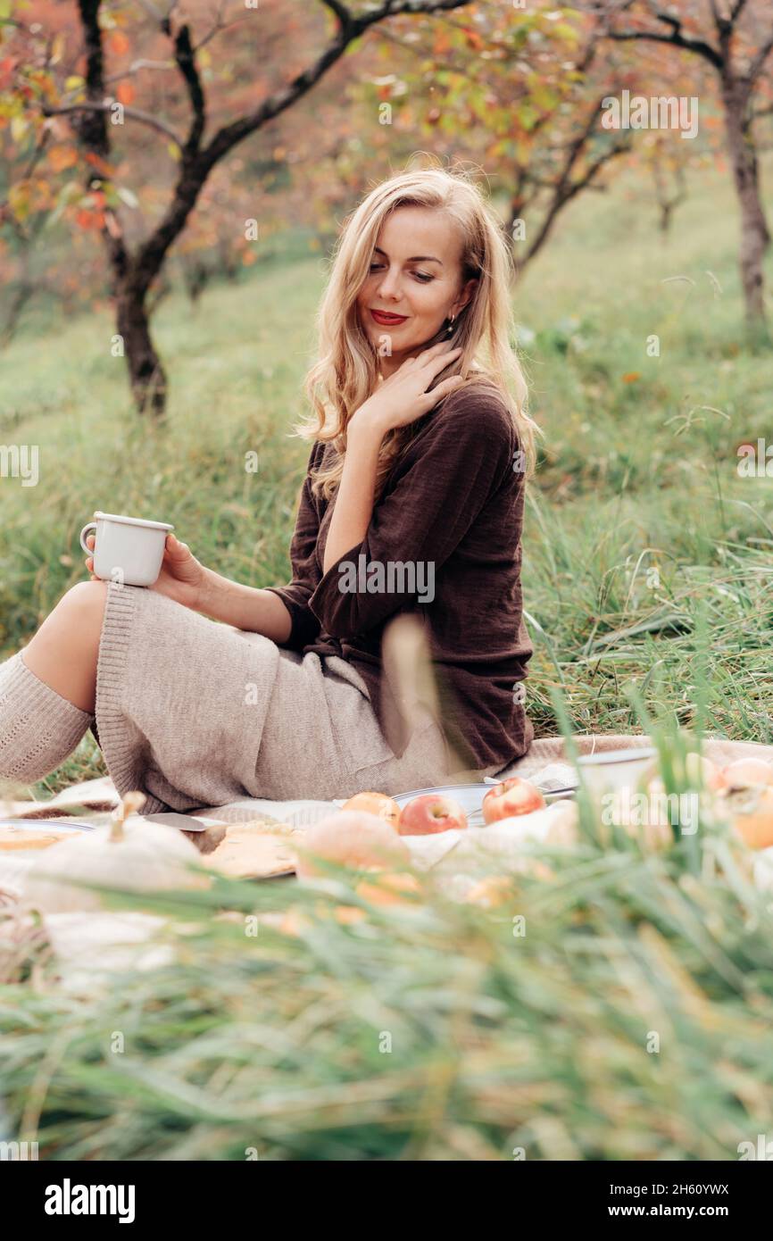Vertikales, ehrliches Porträt einer charmanten Frau, die bei einem Picknick im Herbstgarten sitzt. Stockfoto