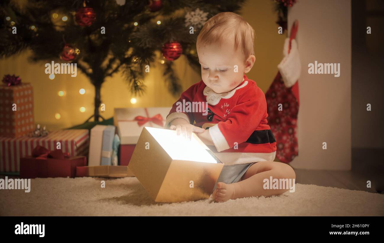 Niedlicher Baby Junge im Weihnachtsmann Kostüm, der im Weihnachtsgeschenk-Kasten schaut. Familien und Kinder feiern Winterferien. Stockfoto