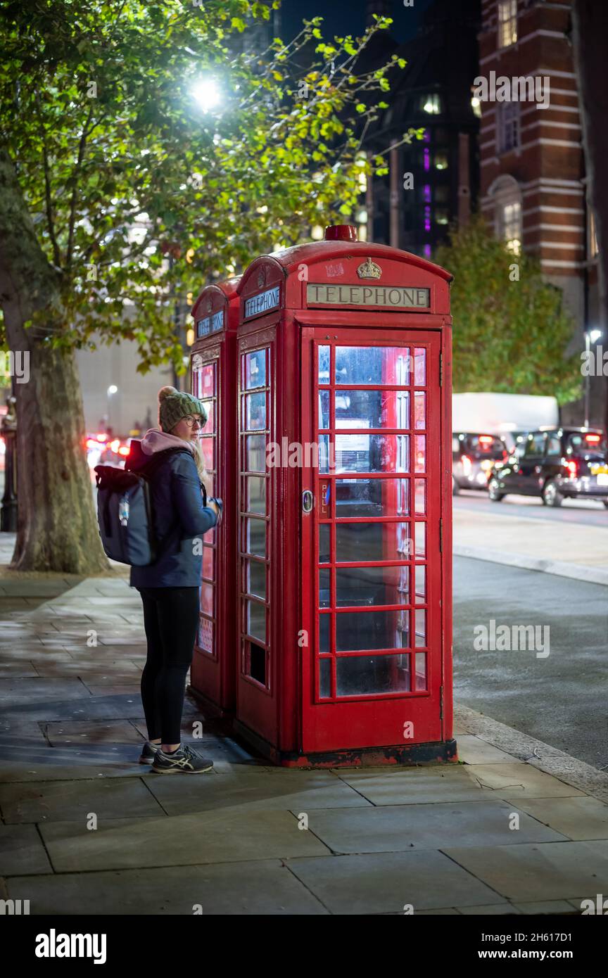 LONDON - 1. NOVEMBER 2021: Eine junge Frau mit Rucksack außerhalb der traditionellen britischen roten Telefonzellen in der Nacht Stockfoto